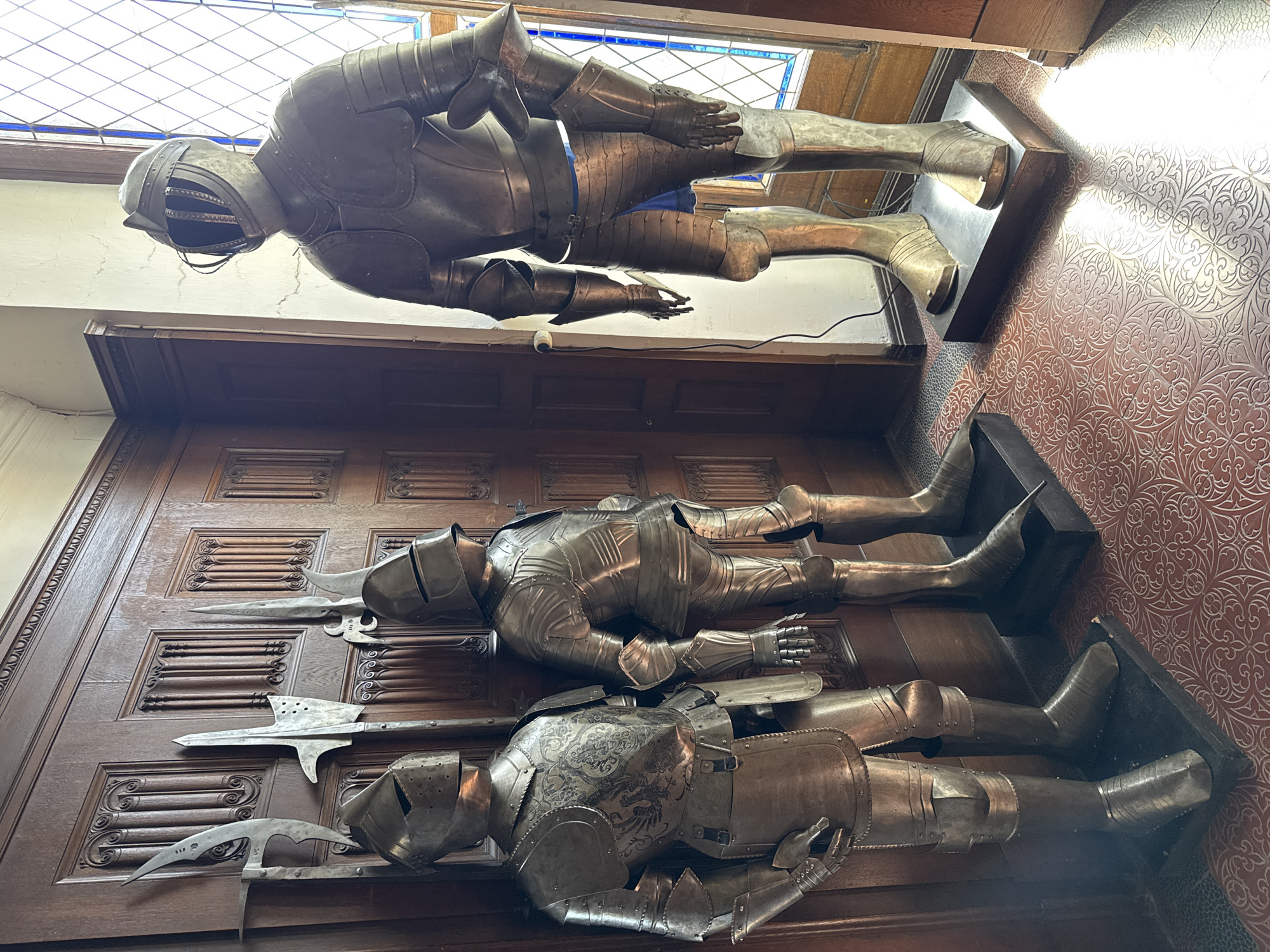 The image depicts three full suits of armor displayed at Reichsburg Cochem, a castle located in Cochem, Germany. Two suits are mounted on a dark brown wooden wall. Both are displayed lying down. Polearm weapons of medieval design are mounted along with the lower two suits of armor. The third suit is mounted horizontally above them, placed on a wall beneath a window. All three suits appear to be made of polished metal.