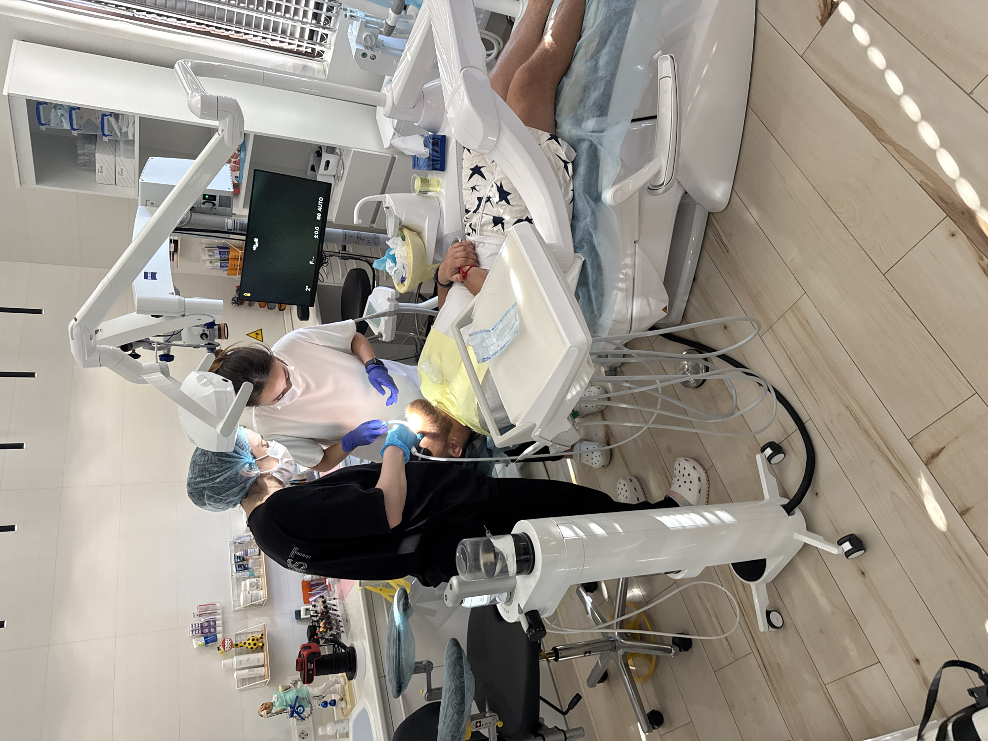 The image depicts a dental examination room, possibly located in Mirnoye, Ukraine, given the context. Two female dental professionals, wearing surgical caps and masks, are attending to a male patient who is reclined in a dental chair. One professional, wearing a black shirt, is positioned on the left, holding dental instruments and appearing to work on the patient's mouth. The other professional, in a white shirt, is on the right, also engaged in the procedure. The patient is lying on a light blue dental drape, wearing patterned shorts.
The room is equipped with standard dental apparatus, including a dental chair, a monitor displaying a green screen with text overlays, and various medical equipment on wheeled stands. Shelves on the wall hold dental supplies and instruments. The floor is made of light-colored wooden planks. The overall environment suggests a clinical setting focused on dental care.
