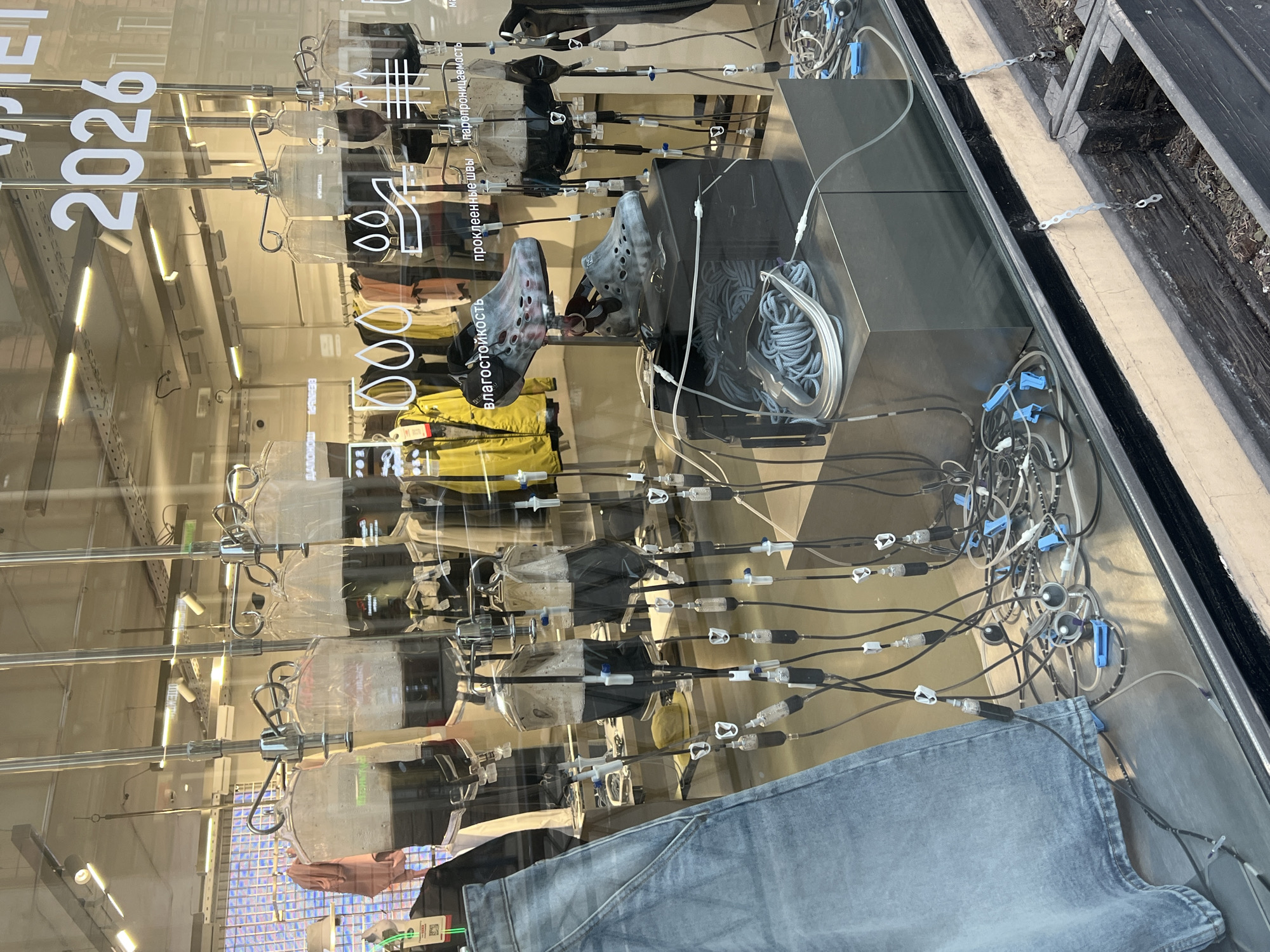 The image captures a unique and conceptual store window display, likely located in Saint Petersburg, Russia. The primary subjects are numerous transparent plastic IV drip bags, each suspended from metal stands, reminiscent of hospital equipment. Thin medical-like tubing extends from these bags, connecting them to various items of clothing and footwear arranged within the display.
Visible items include a pair of gray, speckled, Crocs-like shoes, a neatly coiled gray rope or cord, a pair of blue denim jeans, and several jackets in colors such as yellow, black, and beige/pink. The tubes appear to be "infusing" a clear liquid into these products, creating a visual metaphor. A person's hand is partially visible at the bottom left, seemingly holding or interacting with an item near the jeans, potentially part of the display arrangement.
The background, seen through the clear window, reveals the interior of a retail store with clothing racks and bright strip lighting. Outside the window, a glimpse of a dark ground surface and some natural debris is visible, indicating an urban setting.
Prominently displayed on the window glass is large white text in Russian. The year "2026" stands out. Other technical terms, likely describing product features, include:
* "влагостойкость" (vlagostoykost), meaning "water resistance," accompanied by a raindrop icon.
* "проклеенные швы" (prokleennye shvy), meaning "taped seams" or "sealed seams," with a corresponding stitch icon.
* "паропроницаемость" (paropronitsaemost), meaning "vapor permeability" or "breathability," depicted with an arrow icon.
The display leverages a medical aesthetic to highlight the advanced or technical qualities of the apparel and footwear, suggesting that features like water resistance, breathability, and sealed seams are essential components or "ingredients" of the products. The scene appears to be set during the daytime, evidenced by the reflections on the glass. The overall tone is one of innovative and thought-provoking marketing.