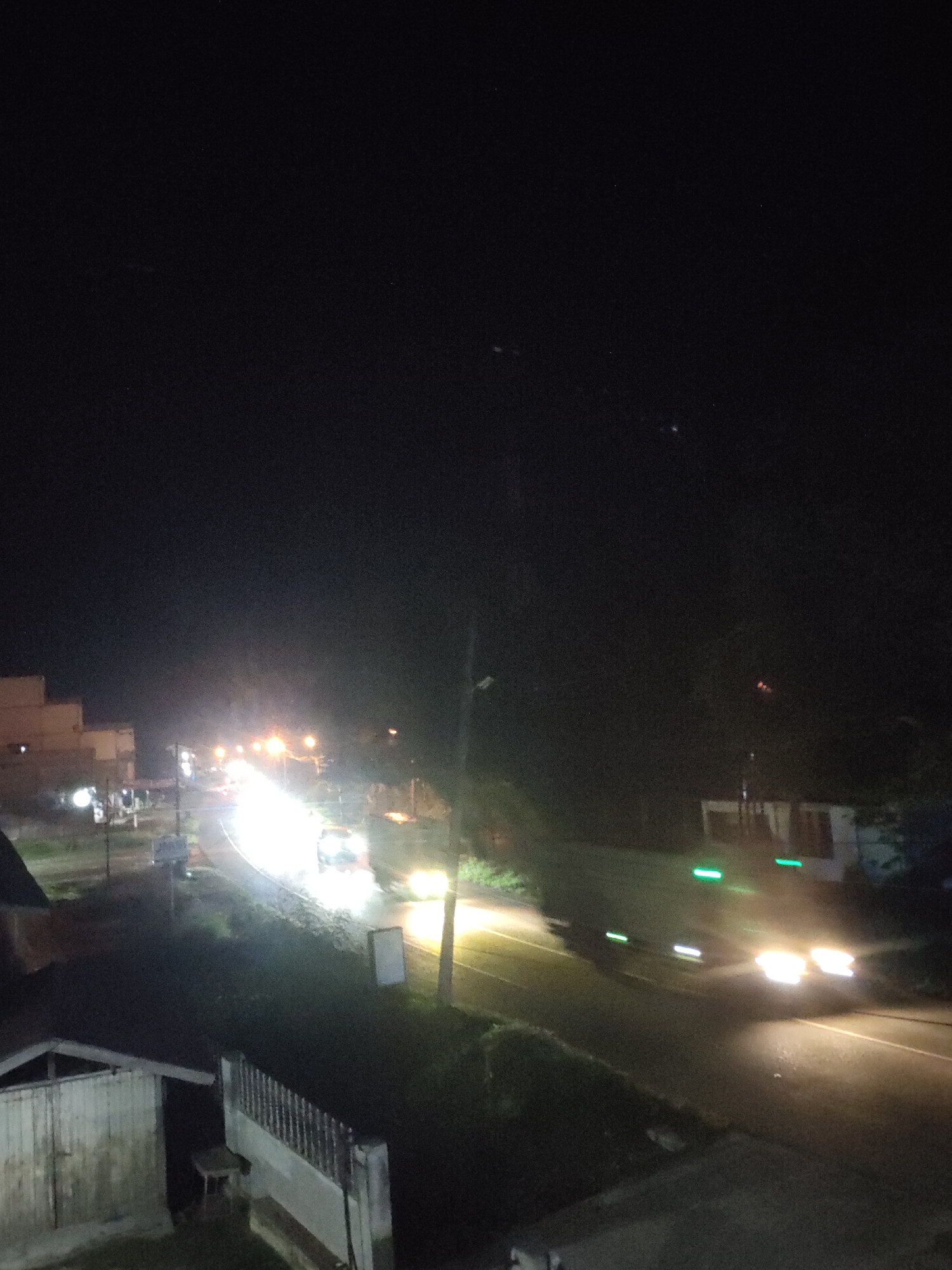 The image captures a nighttime scene on a road in Meunasahaluedrienlhoksukon Barat, Indonesia. A street or road curves through the frame, illuminated by the headlights of passing vehicles and streetlights. Several cars are visible, with their lights creating streaks due to motion blur, indicating they are in transit. Buildings are present on the left side of the road, with some showing lights within. The overall atmosphere is dark, typical of a rural or suburban area at night, with the artificial lights providing the primary illumination. The foreground shows what appears to be a low structure, possibly a shed or a small building, and a fence or railing. Vegetation is visible along the sides of the road and in the background, appearing as dark masses against the night sky. The presence of streetlights and moving vehicles suggests a functional road system within the locality.