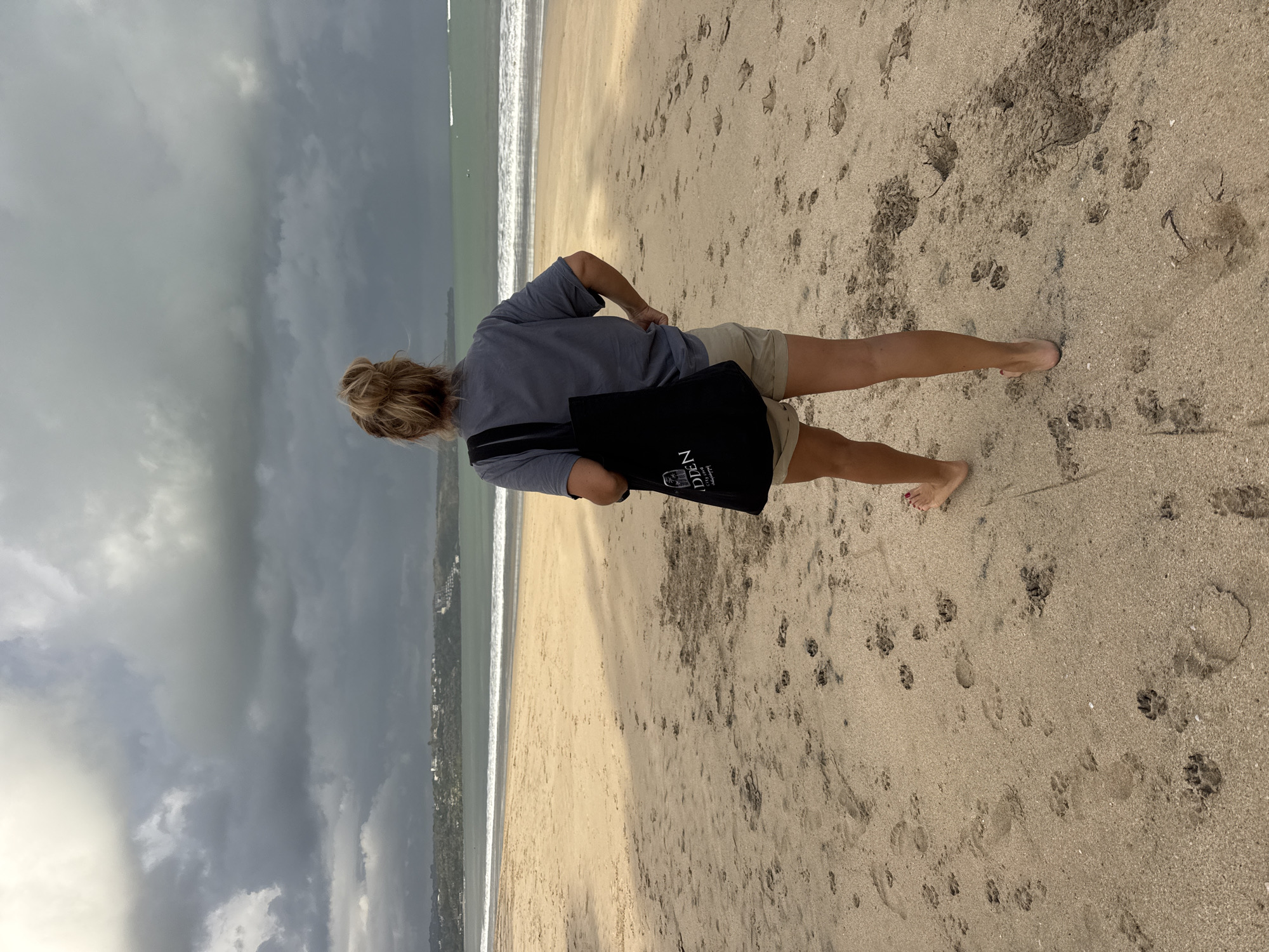 In Kuta, Indonesia, a woman stands on a sandy beach, facing the ocean. The sky is overcast with dark clouds, suggesting approaching rain. The water is a murky green, with moderate waves breaking on the shore. The beach is covered in footprints and paw prints, indicating recent activity. The woman is wearing a gray t-shirt and tan shorts, carrying a black tote bag with a white logo. She is barefoot, with her left hand resting on her hip. In the distance, a line of buildings and greenery is visible along the coast.