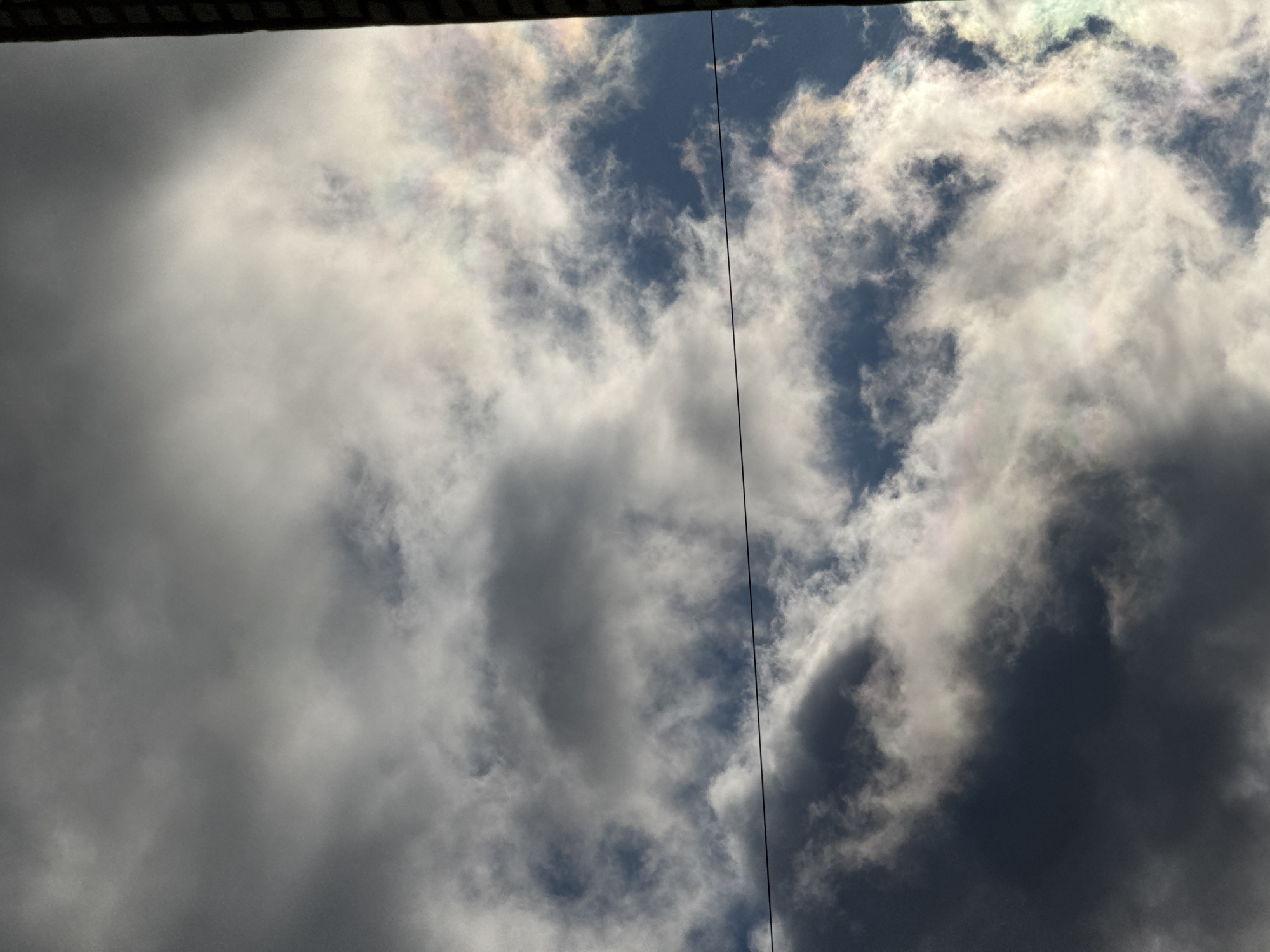 The image captures an upward view of a cloudy sky, dominated by various cloud formations against a backdrop of patches of clear blue sky. A distinct, thin dark line, likely a utility wire or cable, runs vertically down the center-right of the frame. At the very top edge, a dark, horizontal, and slightly textured band, possibly the underside of a roof or an architectural overhang, is visible.
The clouds themselves vary in density and brightness. On the left and bottom right, there are heavy, dark grey clouds, suggesting considerable thickness or shadow. In contrast, many areas feature bright white clouds, particularly towards the right and upper-middle sections. A notable detail is the presence of cloud iridescence (irisation), visible as subtle rainbow-like colors—hints of green, pink, and yellow—on the edges and within some of the thinner, brighter clouds, especially towards the upper right. This phenomenon indicates the diffraction of sunlight by small water droplets or ice crystals in the clouds.
The scene depicts a natural sky view during daytime, with no human subjects or explicit activity. The weather appears partly cloudy to overcast. No text is visible in the image. The context provided is Chaikovsky, Russia, although no specific landmarks within the image confirm this location.