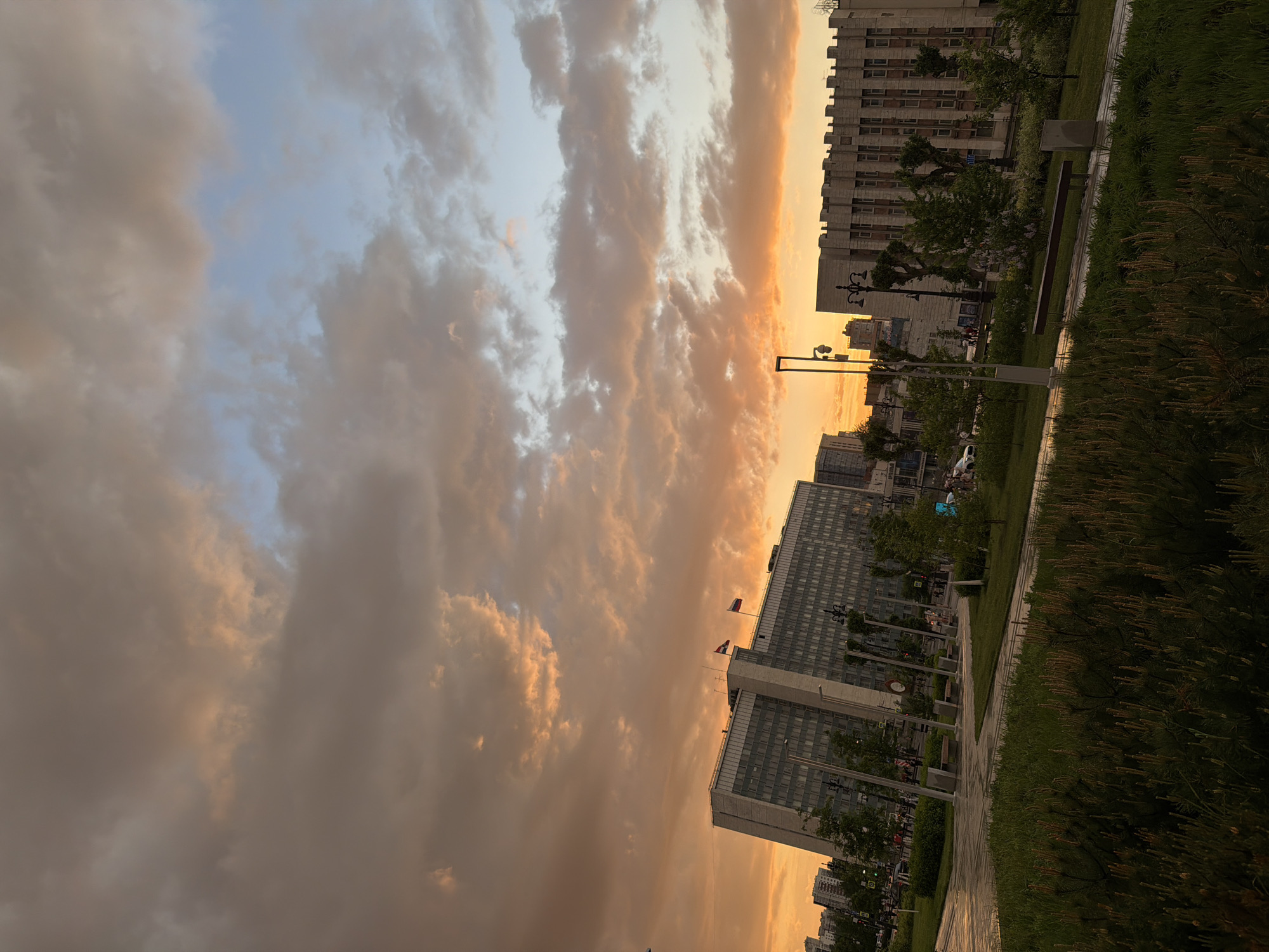 The image shows a cityscape at dusk, likely in Perm, Russia. Dominating the view are large, multi-story buildings with numerous windows. The buildings appear modern, made of concrete and glass. The sky is filled with clouds, illuminated with warm golden and orange hues from the setting sun. The city park stretches along a pedestrian path. A sidewalk runs parallel to green spaces. The scene includes streetlights. The image seems to have been taken from a low angle, looking up towards the buildings and the sky. The scene conveys a sense of urban space with natural elements intertwined.