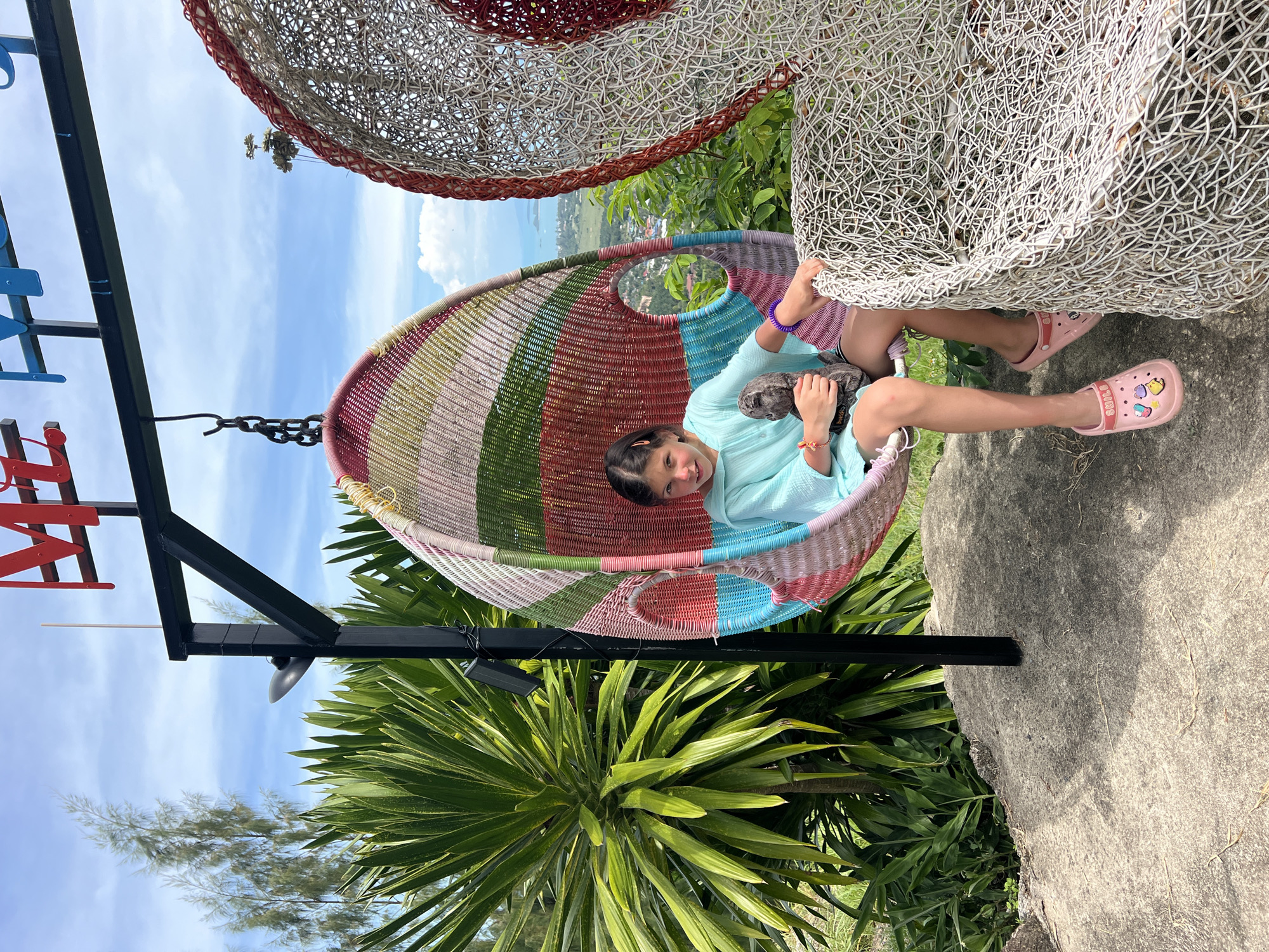A young girl with dark hair is sitting in a colorful woven hanging chair at an outdoor viewpoint in Baan Thurian, Thailand. The chair is suspended from a black metal frame and features stripes of pink, red, green, and blue. The girl is wearing a light blue hooded top and pink clogs, one of which has cartoon charms. She is holding a dark gray stuffed animal. To the right of the hanging chair is a large, white, basket-like sculpture. Behind the girl, lush green tropical plants and trees are visible. In the distance, a town with buildings and a body of water can be seen under a partly cloudy sky. The ground in the foreground is a rough, gray concrete surface.