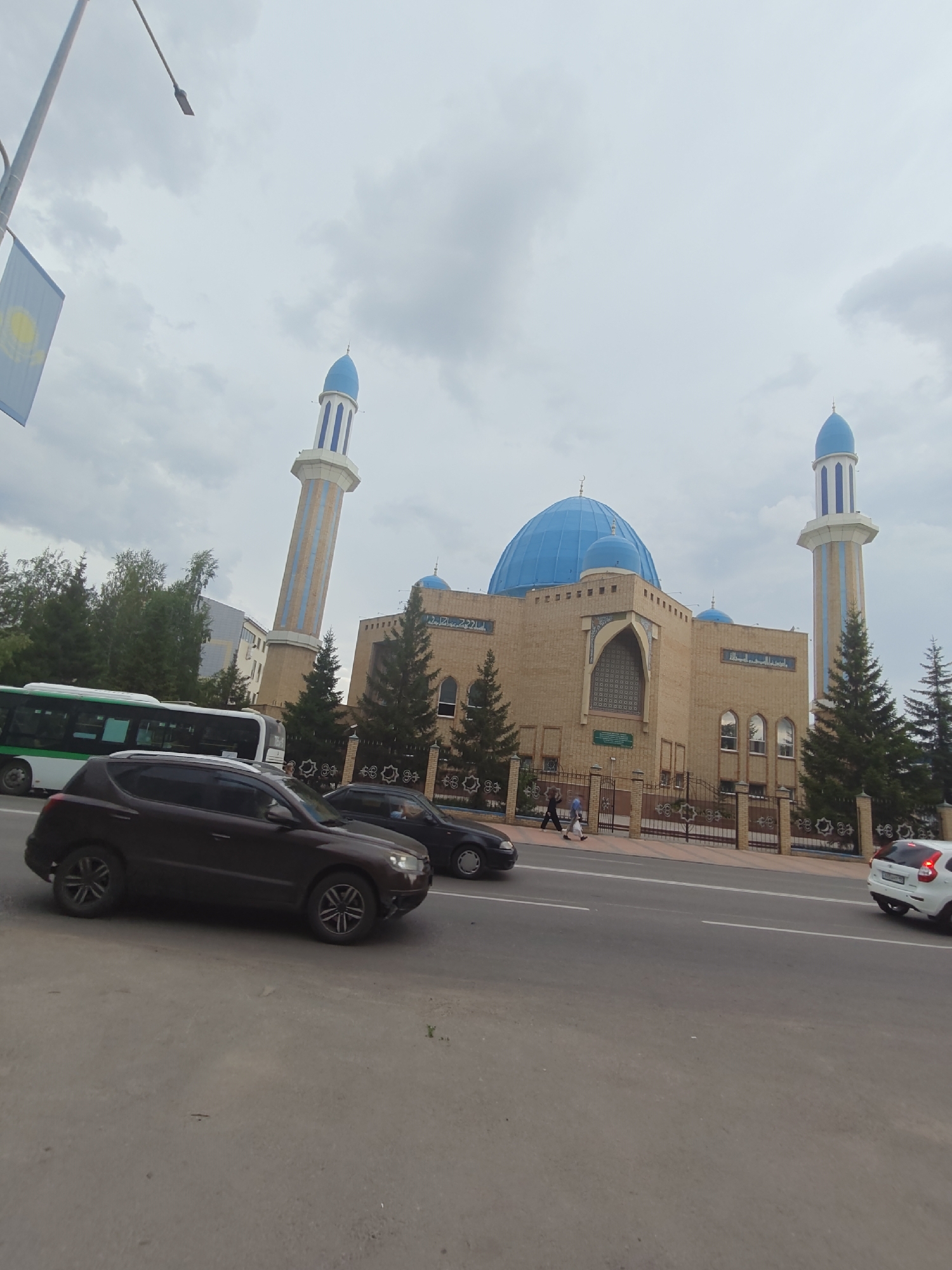 The image shows a mosque with a large blue dome and two tall minarets. The mosque is made of light brown brick and has decorative elements around the windows and doorways. There is a fence in front of the mosque with intricate metalwork.
In front of the mosque, there is a road with several cars and a bus. The cars appear to be driving on the road, and the bus is stopped or moving slowly. There are also a few people walking on the sidewalk in front of the mosque.
The sky is overcast with clouds. The scene appears to be in an urban area, with trees and buildings visible in the background. The image is taken from a low angle, looking up at the mosque.
Based on the presence of the flag of Kazakhstan and the architectural style of the mosque, the location is likely the Central Mosque of Petropavl, Kazakhstan. The mosque's unique architectural features, such as the blue dome and the style of the minarets, are characteristic of this mosque. The surrounding environment with trees, a road, and a bus further supports this identification of the location in the city of Petropavl.