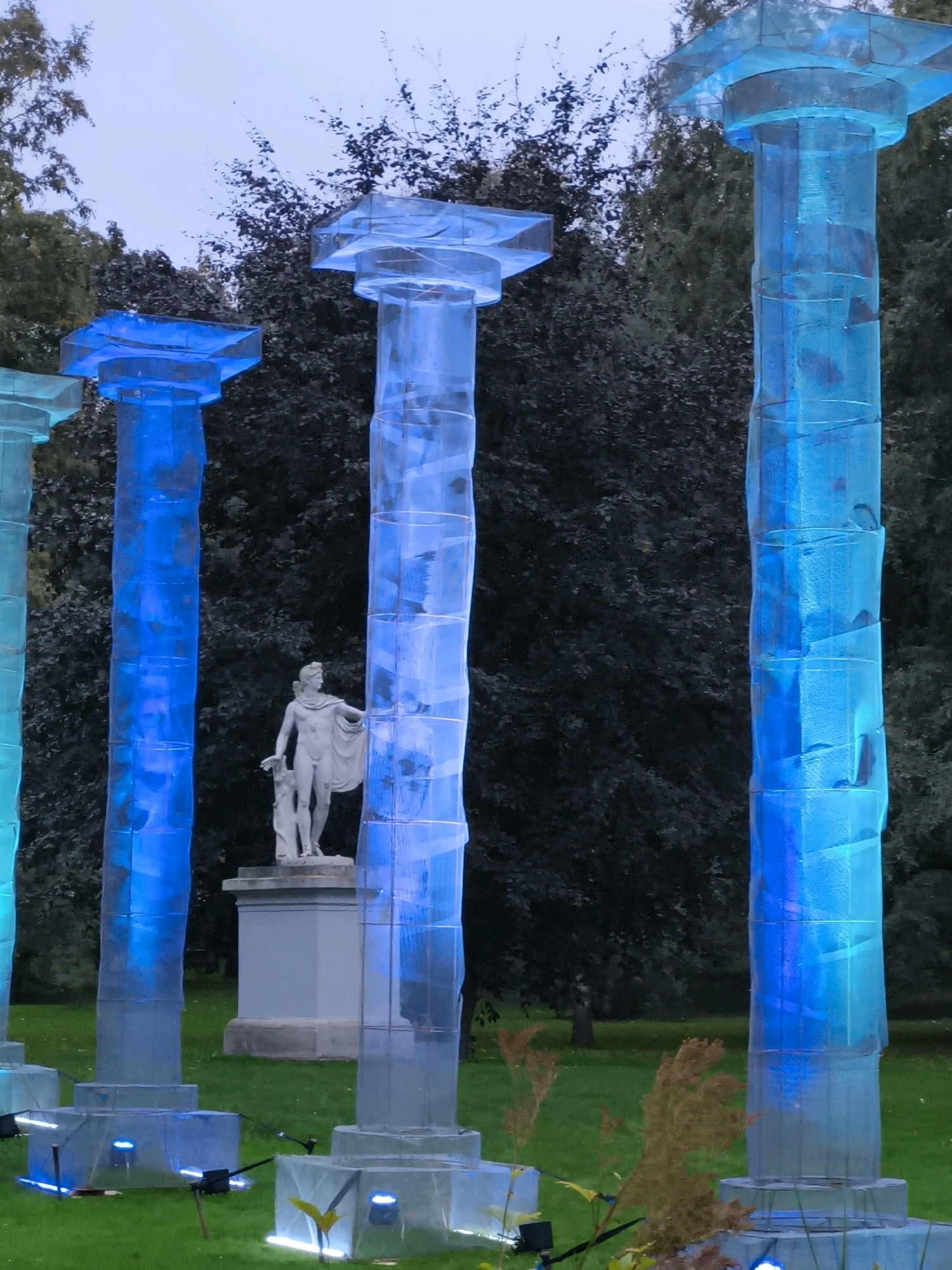 The image depicts an outdoor art installation featuring several tall, translucent pillars illuminated with blue light, resembling futuristic columns. These structures are arranged on a grassy lawn in front of a dark, dense backdrop of trees. In the middle ground, a white classical statue of a male figure, possibly a god or hero, stands on a pedestal. The statue is partially obscured by the illuminated pillars. The pillars appear to be made of a mesh-like material wrapped around a cylindrical structure, creating a textured surface that catches and diffuses the blue light. The base of each pillar is also illuminated with white lights. The scene suggests a nighttime or twilight setting due to the artificial lighting and the dim natural light. The location is identified as Tallinn, Estonia.