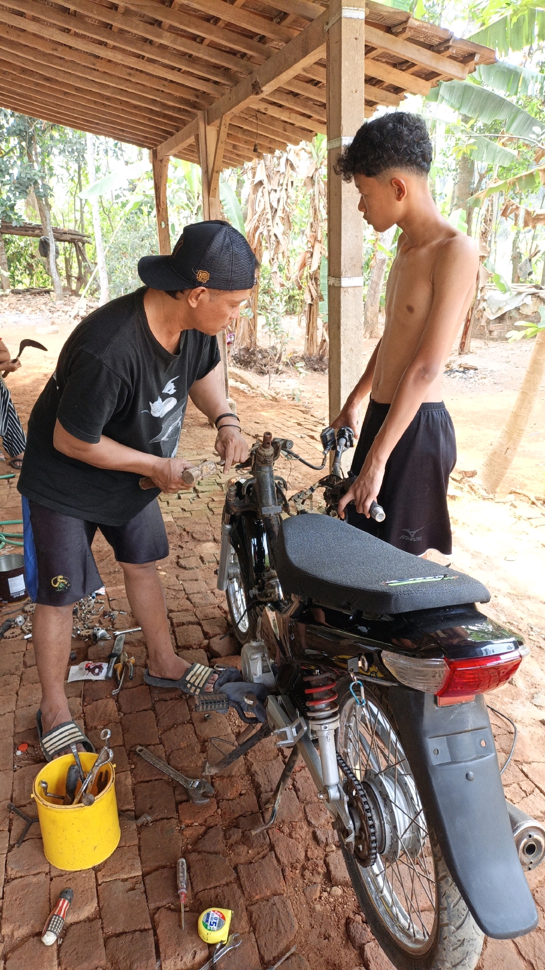 In Suwawal Timur, Indonesia, a man wearing a black t-shirt, black shorts with blue trim, and a black baseball cap is seen working on a black motorcycle. He is holding a tool and appears to be adjusting a part of the motorcycle's handlebars. To his right, a shirtless young man with curly hair, wearing black shorts, stands beside the motorcycle, holding onto its handlebars. Tools, including wrenches, a yellow bucket filled with various tools, a screwdriver, and a measuring tape, are scattered on the brick ground around the motorcycle. The scene takes place outdoors under a wooden-covered structure, with trees and foliage visible in the background. The ground is made of reddish-brown bricks.
