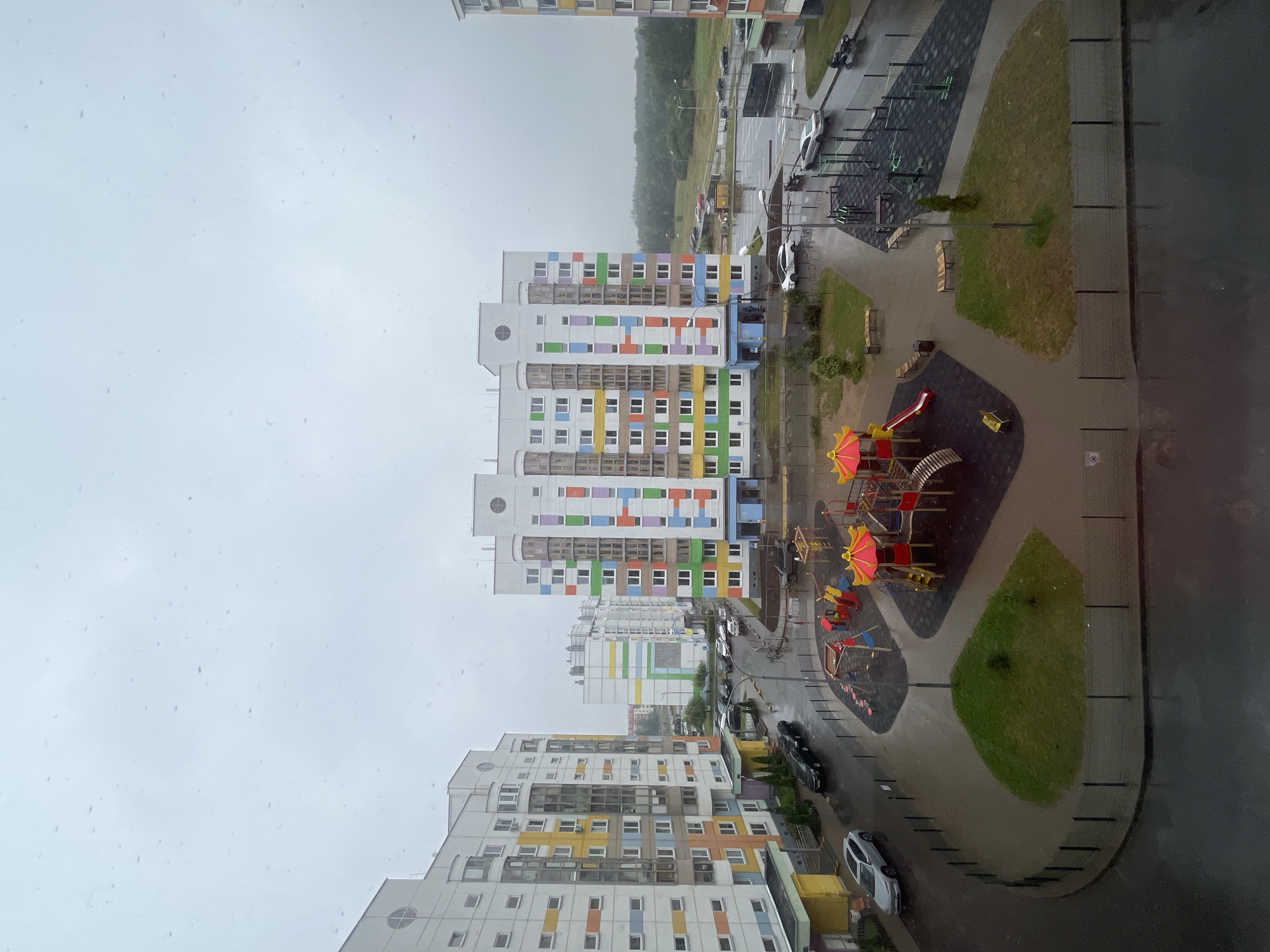 The image depicts an outdoor view of a residential courtyard in a modern housing complex in the Senitsa village council, Belarus. The scene is overcast and appears to be raining, with visible raindrops on the camera lens. In the foreground, a colorful playground with slides, swings, and climbing structures is visible. The ground around the playground is paved with dark rubber and light-colored pavers. A small grassy mound with a tree is situated nearby, enclosed by a dark metal fence.
Several multi-story apartment buildings with distinct architectural styles and colorful facades dominate the background. These buildings feature a pattern of brightly colored rectangular panels in shades of yellow, orange, green, blue, and purple against white exteriors. Cars are parked on the wet asphalt road winding through the complex, and a few are in motion. Beyond the buildings, a treeline and a flat, green landscape suggest the suburban or rural outskirts of the area. The overall impression is one of a recently developed, well-maintained residential area in a damp, overcast environment.