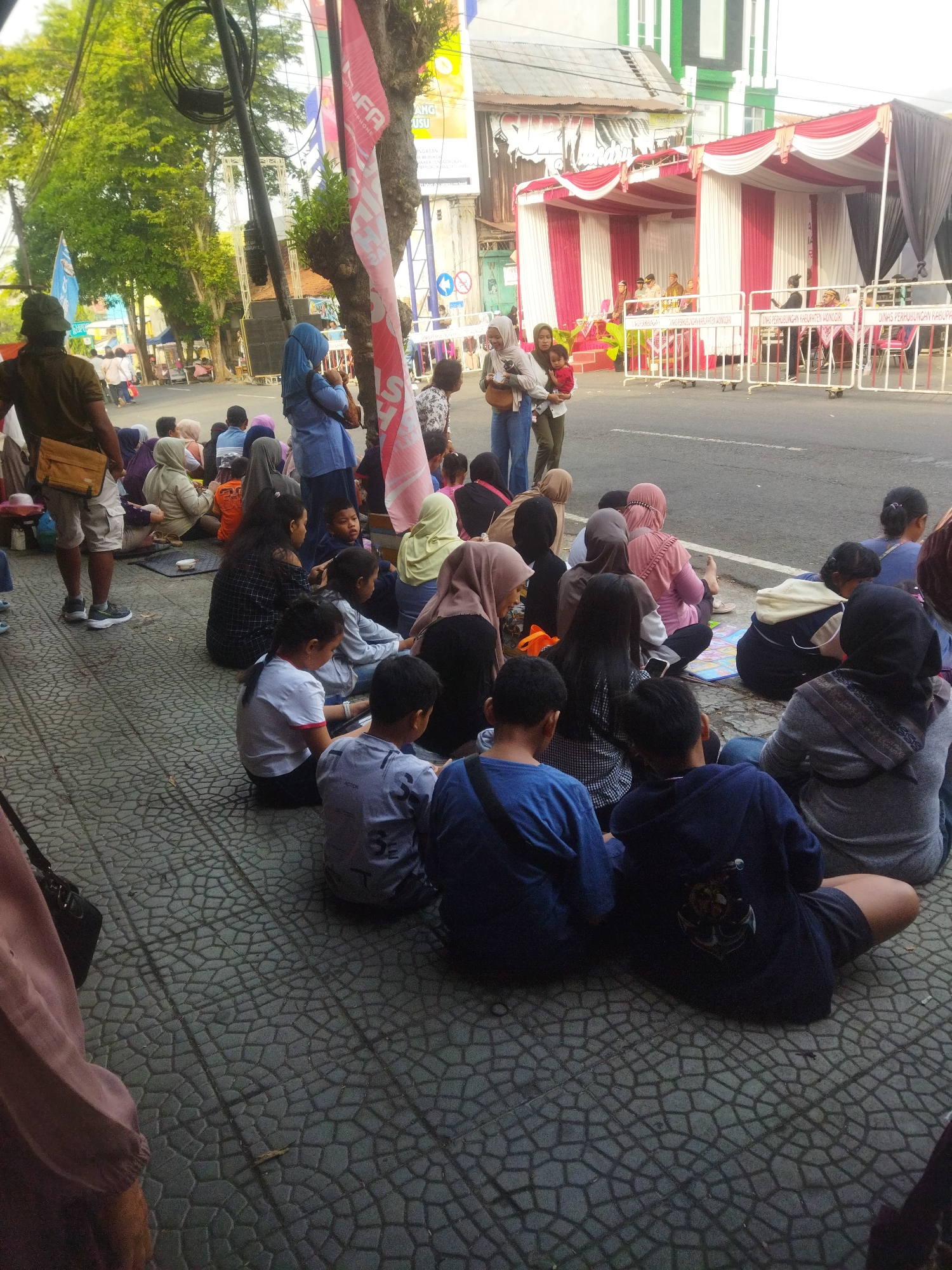 The image captures a scene on a street in Wonogiri, Indonesia, likely during an event given the presence of a temporary stage with red and white draped fabric. A crowd of people, predominantly women and children, are seated on a paved sidewalk facing the street. Many are wearing hijabs, indicating a Muslim-majority population.
In the foreground, several children and adults are sitting cross-legged on the ground. A man in a dark blue hoodie with an emblem on the back is prominently featured. To his left, a person in a grey hoodie and a person in a grey t-shirt are also seated. Further to the left, a child in a grey t-shirt is visible.
In the middle ground, a diverse group of individuals are seated, some looking towards the street. A woman in a blue hijab and a woman in a light green hijab are among them. Behind them, a few people are standing, and further back, there are tents and banners, suggesting an organized event. A large black speaker is mounted on a pole.
In the background, the street is visible with people walking and some vehicles. The buildings lining the street appear to be a mix of commercial and residential structures, with some displaying vibrant colors. The overall atmosphere suggests a public gathering, possibly a festival, performance, or community event, taking place in a bustling urban environment. The lighting indicates it is daytime, likely morning or afternoon, given the soft natural light.