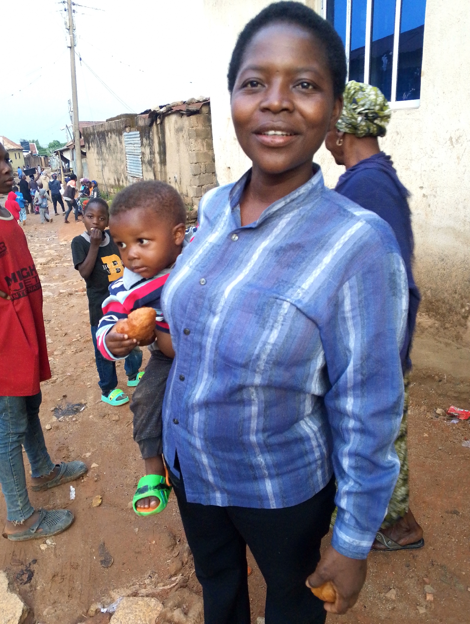 The image depicts an African woman holding a toddler in front of a weathered building with a blue-framed window in a district of Jos, Nigeria. The woman is wearing a blue striped shirt and black pants. The child is wearing a striped shirt, grey pants, and green sandals, holding what appears to be a fried dough ball. Other children and adults are visible in the background on a dirt road with scattered debris. A power pole is visible in the background. The ground is red clay.