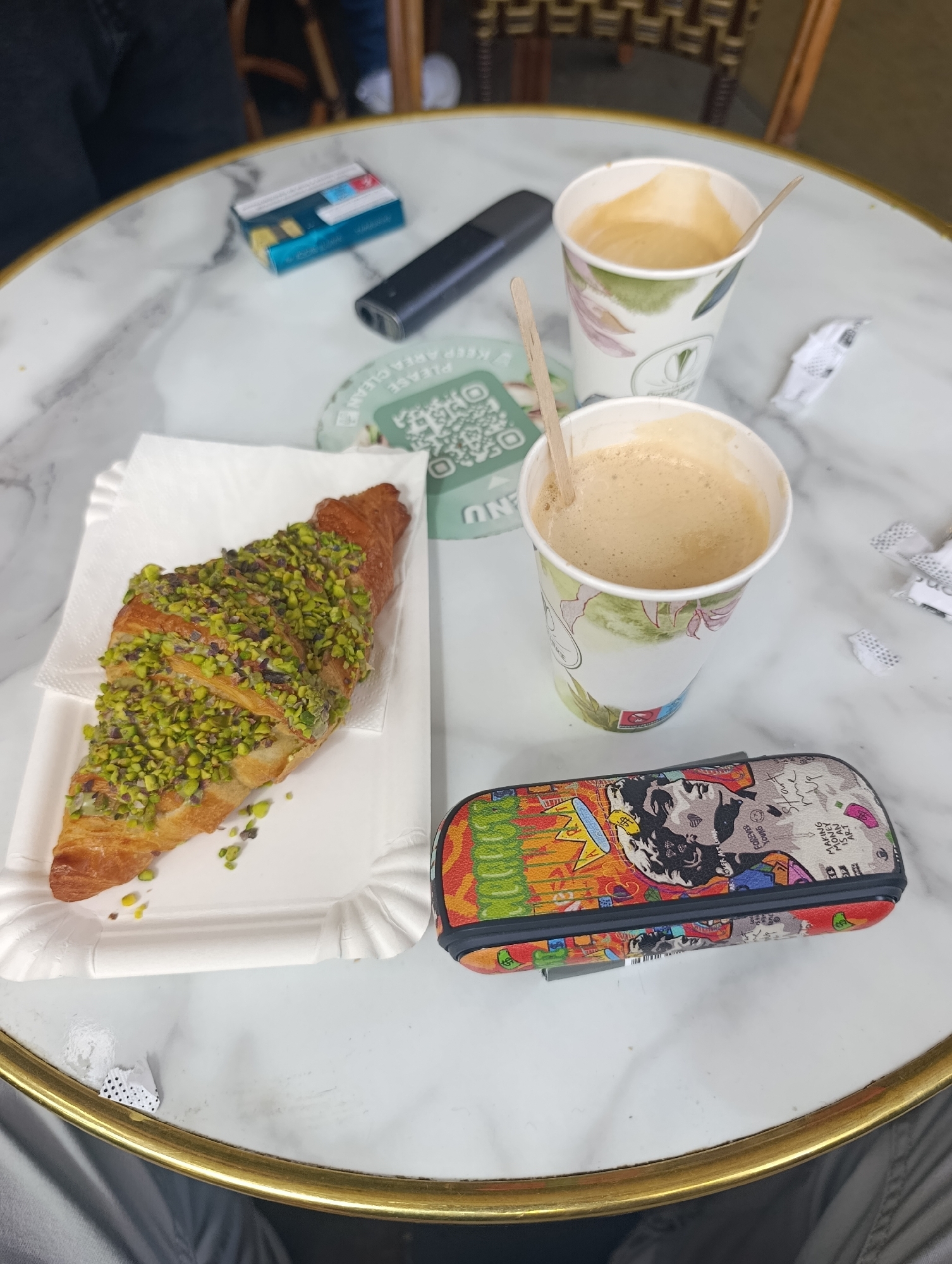The scene depicts a marble table with a gold rim, likely in a Parisian cafe in France. A croissant topped with chopped pistachios sits on a white paper plate. Two paper cups containing coffee with foam are present, each with a wooden stir stick. A pack of cigarettes, a vaping device, a QR code sign, a pencil case with a colorful pop art design, and various pieces of discarded paper are scattered on the table. Two individuals are partially visible, one wearing a dark-colored jacket and the other wearing light-colored pants and shoes.