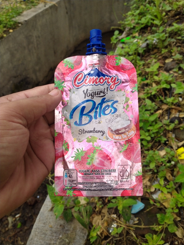A hand holds a pink Cimory Yogurt Bites pouch with a blue spout, featuring a strawberry flavor with nata de coco. The pouch, which appears slightly worn, is held against a bright outdoor background of green vegetation and a concrete structure.
Visible text on the pouch includes:
- "Cimory Yogurt! Bites"
- "Strawberry ~ with Nata De Coco"
- "YOGURT RASA STROBERI DENGAN NATA DE COCO" (Strawberry flavored yogurt with nata de coco)
- A recommendation to keep it at "4°C"
- "BERAT BERSIH / NET WEIGHT: 120g"
- "HALAL INDONESIA" logo
- "BPOM RI MD 242811001000/10"
- Partially visible text: "DIPRODUKSI OLEH / MANUFACTURED BY PT. Cisarua Mountain Dairy, Tbk. Kab. Bogor 16830 - Indonesia"
The setting is outdoors, likely during the daytime, with ample natural light. The background features overgrown green plants and a concrete barrier or wall, suggesting an informal outdoor environment. This image was captured in Karanganyar, Indonesia.