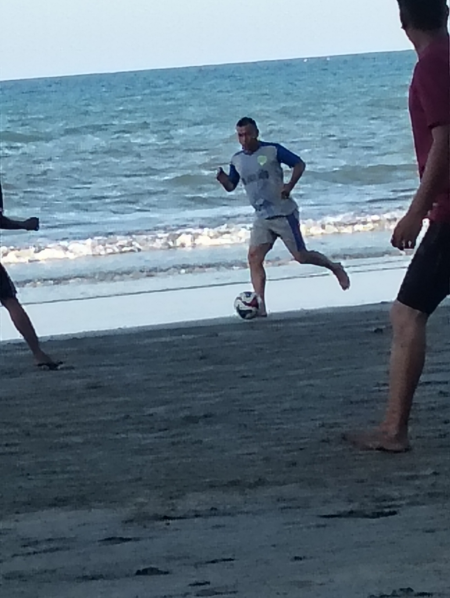 Three individuals are visible playing soccer on a sandy beach next to the ocean. The person in the center, wearing a light blue and white soccer jersey and grey shorts, is kicking a soccer ball with their right foot. Their left arm is bent at the elbow, and their body is angled towards the viewer. To the right, another person, facing away from the camera, is wearing a dark red t-shirt and black shorts. Their right arm is bent, and they are standing with their legs apart. To the left, a third person, mostly out of frame, is wearing dark shorts and is visible from the knees down, with their left leg extended. The ocean is in the background, with gentle waves breaking on the shore. The sky is a pale blue. The location is the beach in Peukan Sot, Indonesia.
