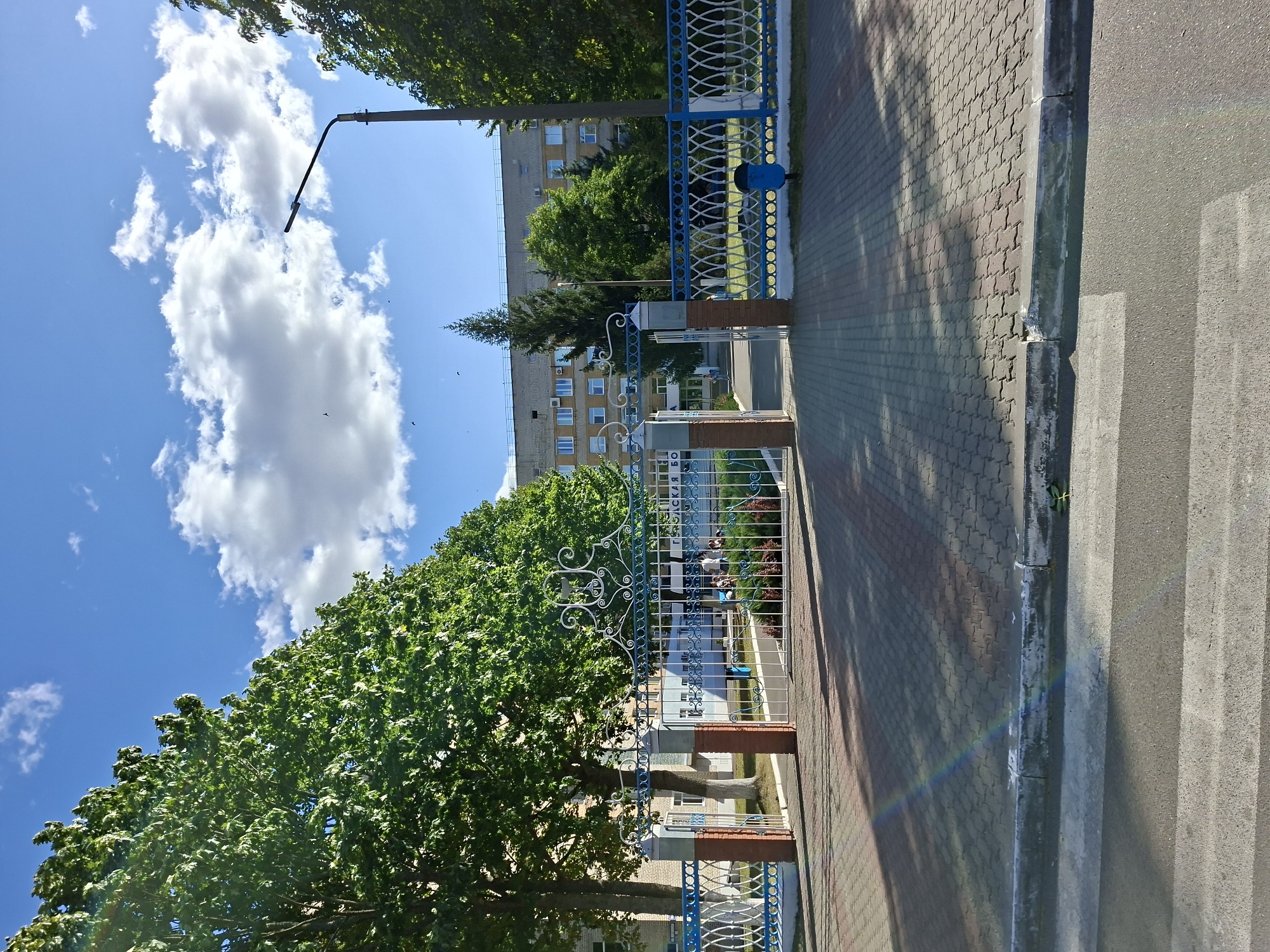 The image captures the entrance to a hospital in Mazyr, Belarus. A white decorative metal gate with blue accents stands open, revealing a paved courtyard and a multi-story building in the background. The building appears to be a brick structure with numerous windows. Lush green trees and a tall pine tree surround the entrance and the building. A lamppost stands to the right of the gate. The foreground shows a crosswalk and a sidewalk paved with interlocking bricks, with shadows from the trees cast across it. The sky is blue with scattered white clouds, and a faint rainbow effect is visible in the lower left of the image. Several people, appearing to be children, are gathered in the courtyard inside the gate, and a few more figures are visible near the building entrance. A blue trash bin is situated near the fence to the right. The overall scene suggests a bright, sunny day.
