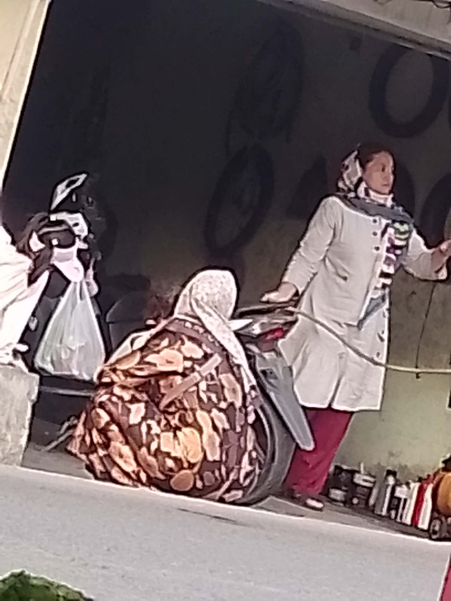 In Cikalongkulon, Indonesia, two individuals are visible in an outdoor setting, possibly a workshop or storage area with a dark background featuring circular motifs. On the left, a person is partially obscured, holding a light-colored plastic bag. To the right, a woman wearing a light-colored tunic and a patterned headscarf stands next to a motorcycle. She is wearing red pants and has her left hand resting on the motorcycle. Another person, also wearing a patterned headscarf and a patterned garment, is seated on the ground behind the motorcycle. The ground surface appears to be paved.