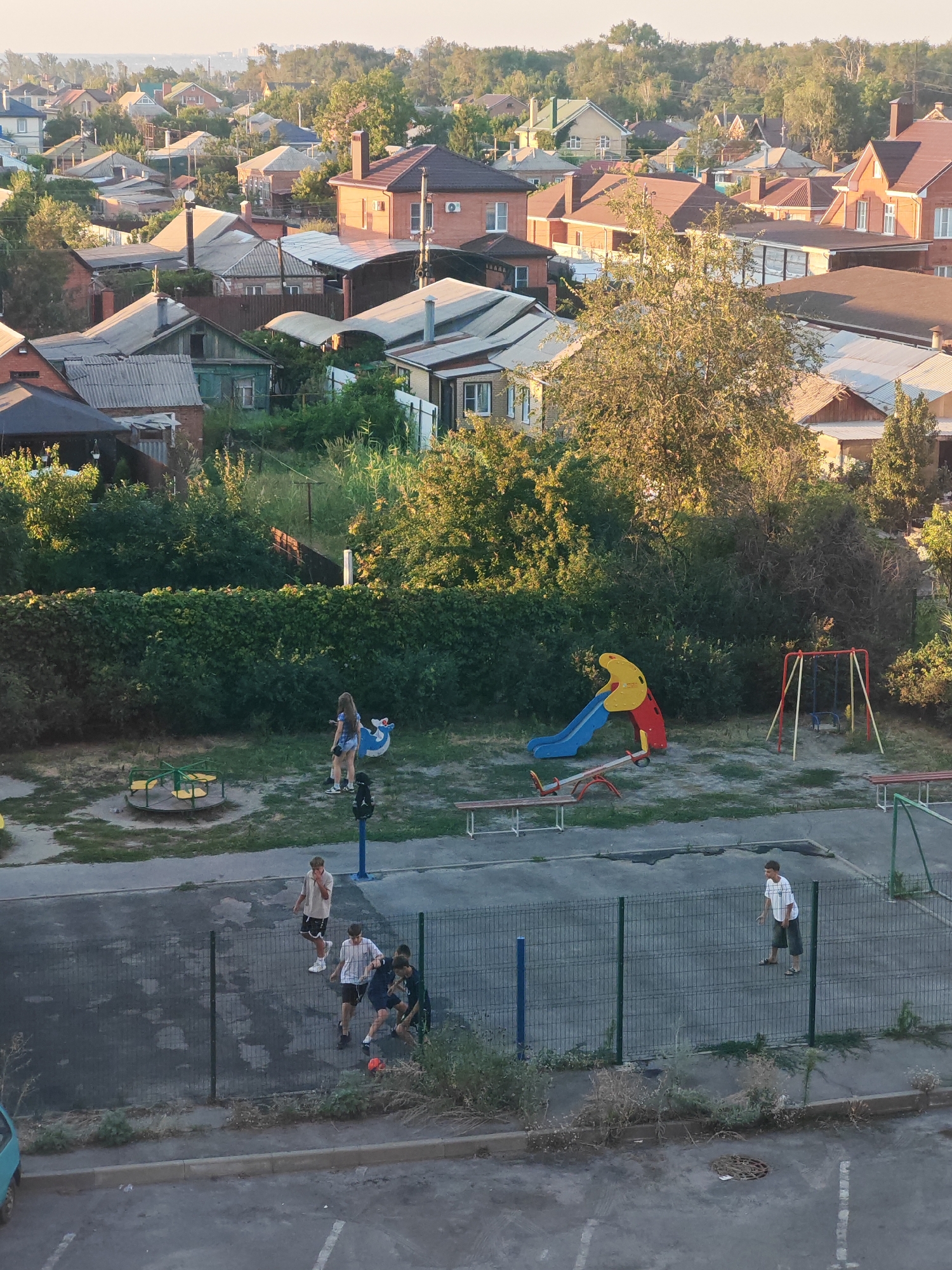 An aerial view captures a residential area in Bataysk, Russia, with numerous houses featuring red-tiled roofs and varying architectural styles. The scene includes dense greenery and trees interspersed among the buildings. In the foreground, a playground area is visible with a slide, swings, a seesaw, and a merry-go-round. Several individuals are present: two young women are near the merry-go-round, and a group of young men are playing soccer on a fenced-in court. Another young man stands on the edge of the court. The ground is a mix of asphalt and patchy grass, with a light blue car parked on the side. The lighting suggests it is late afternoon or early evening.