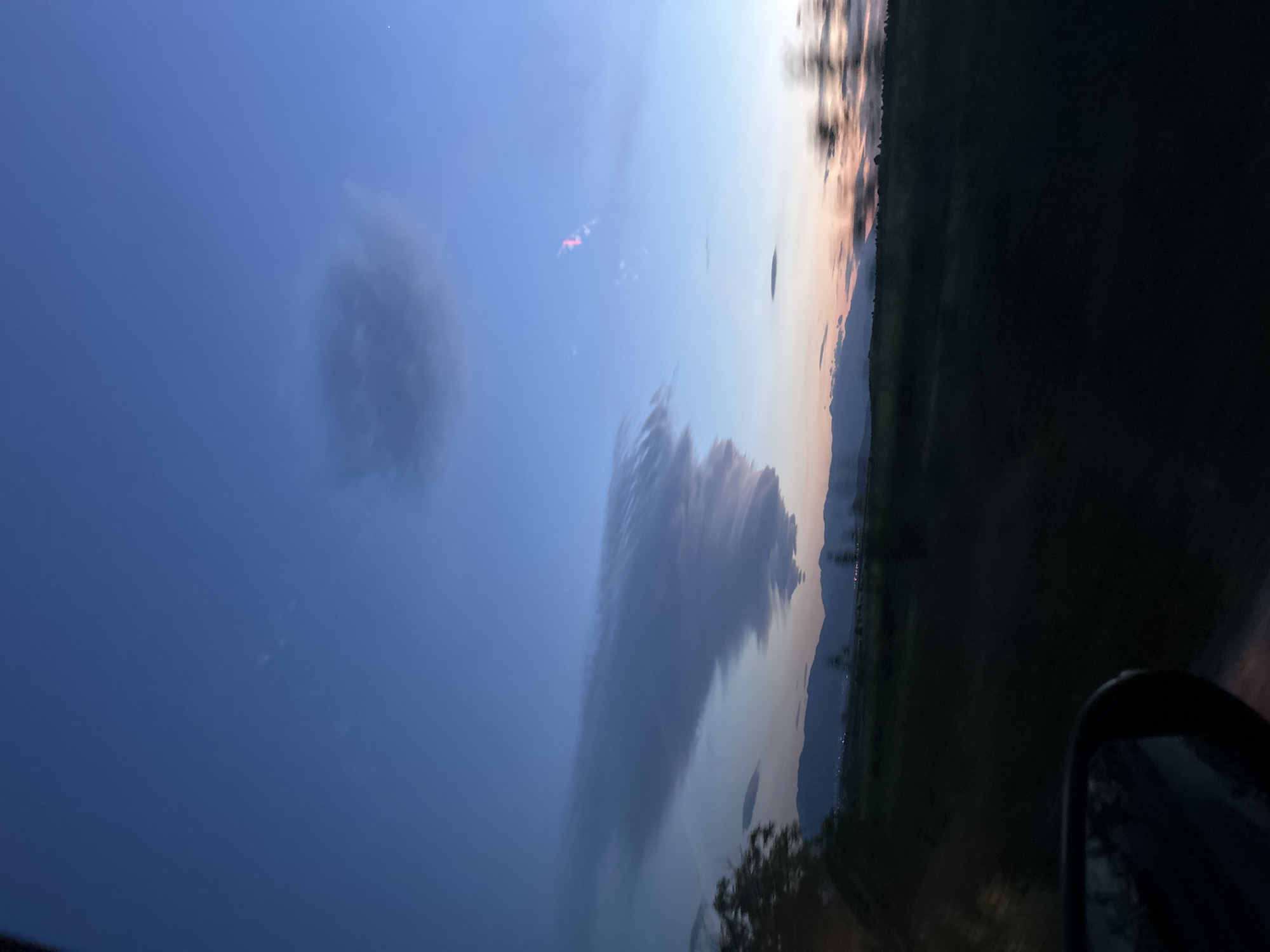 The image depicts a twilight scene, possibly near Ovchepoltsi, Bulgaria. The sky is predominantly blue, with dark clouds forming a long, horizontal shape and a smaller, rounder shape to the left. The horizon shows a transition from light to dark, suggesting a setting sun. The bottom right corner of the image shows the side mirror of a car, indicating the image was likely taken from inside a vehicle. The landscape consists of dark and green vegetation along the horizon. Small lights are visible at the horizon, possibly indicating a town or city.