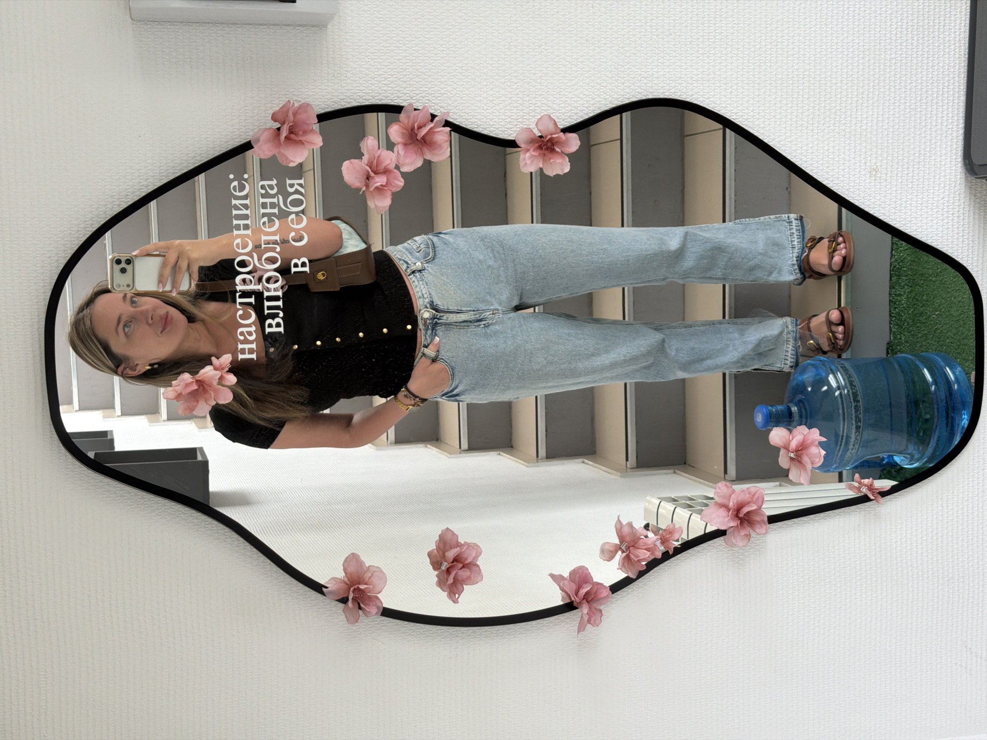 A young woman captures a mirror selfie in Chaikovsky, Russia. The image, originally rotated, shows her standing upright, reflected in a large, heart-shaped or lip-shaped mirror with a black frame. Several artificial pink flowers are attached to the mirror's edges, adding a decorative touch.
The woman has light brown hair and is wearing a dark, possibly black, short-sleeved top with light-wash blue jeans and brown sandals. She is holding a white-cased smartphone to take the picture, with her right arm slightly bent and left arm relaxed. A small brown shoulder bag is worn across her body. Her expression is calm with a slight smile, and she is looking directly at her phone.
The mirror reflects an interior setting. Behind the woman, a grey and white staircase is visible, suggesting a multi-level building. To the right of her legs, a large, blue plastic water cooler bottle rests on what appears to be a green artificial grass mat or carpet. The wall where the mirror hangs is white with a subtle textured pattern.
Prominently displayed in white text on the mirror, near the woman's face, are the Russian words: "настроение: влюблена в себя," which translates to "mood: in love with myself." This text conveys a sense of self-love and contentment, aligning with the woman's relaxed and happy demeanor. The scene suggests a casual moment, possibly in an office or public space, capturing a personal reflection.