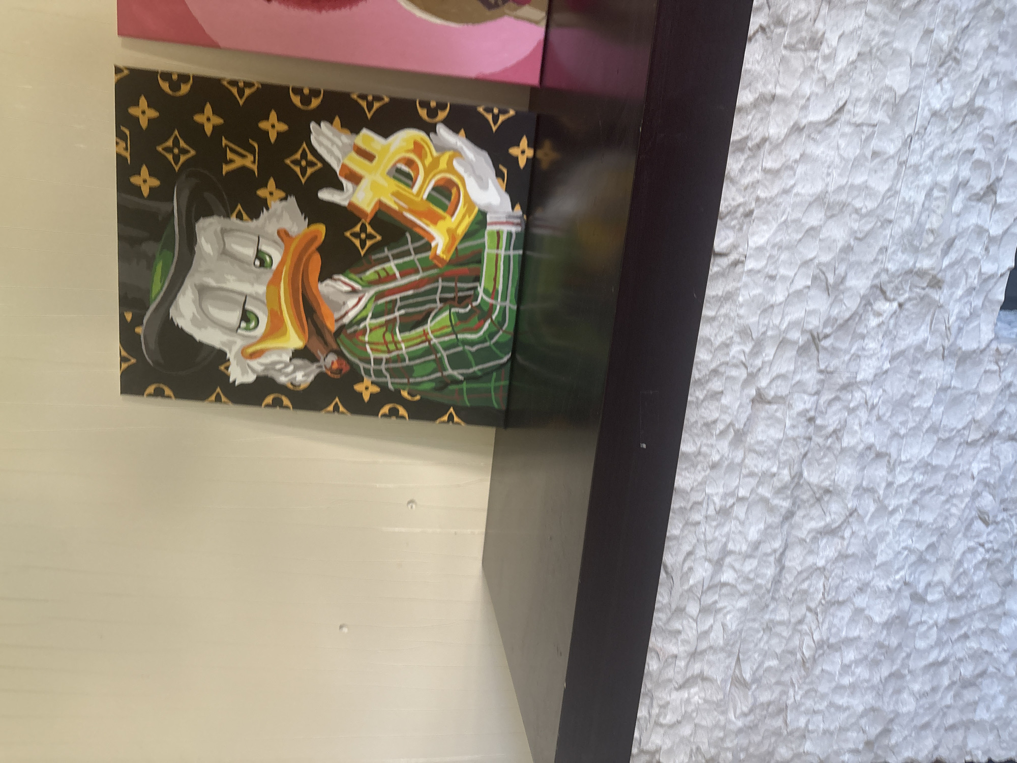 The image shows a painting of Scrooge McDuck wearing a black top hat and green plaid suit, holding a Bitcoin symbol in his gloved hands. The background of the painting features a Louis Vuitton-inspired pattern. The painting is placed on a dark brown shelf, which is mounted on a textured white wall. Another painting is partially visible to the right of the Scrooge McDuck painting. The image does not contain any information about the city of Ореховка, Russia, or its specific location within the city.
