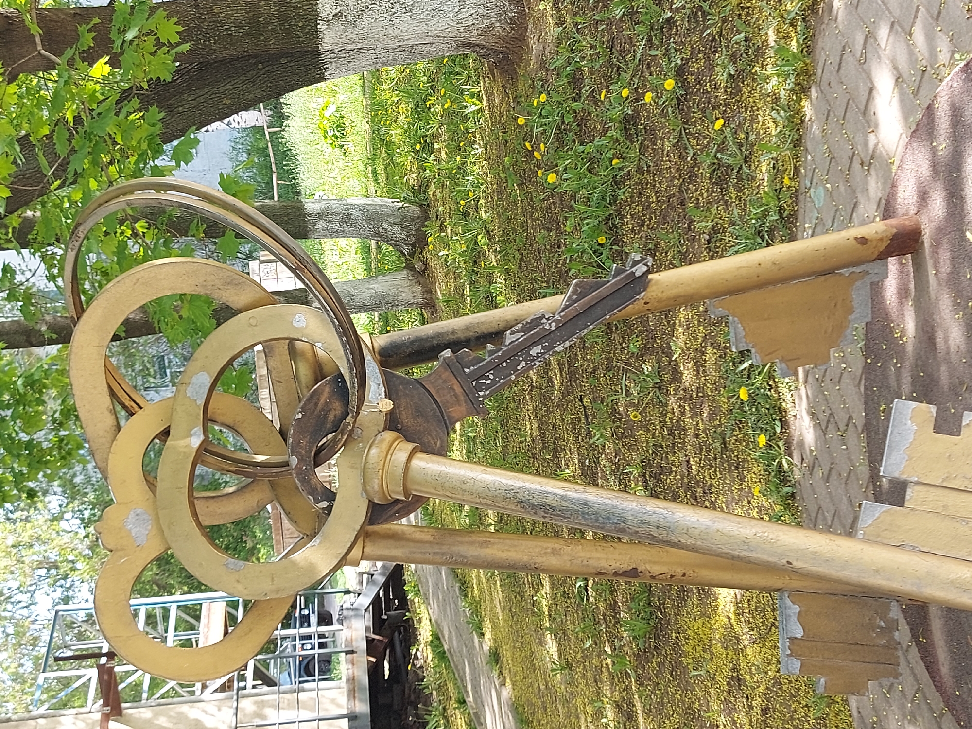 The image depicts a large, decorative set of keys located in Izhevsk, Russia. The keys are metallic and painted in shades of gold and silver. They are positioned on a grassy area with yellow flowers. A tree trunk is visible behind the keys. In the background, there is a fence. Part of a building is also visible behind the fence. The ground consists of brick paving.
