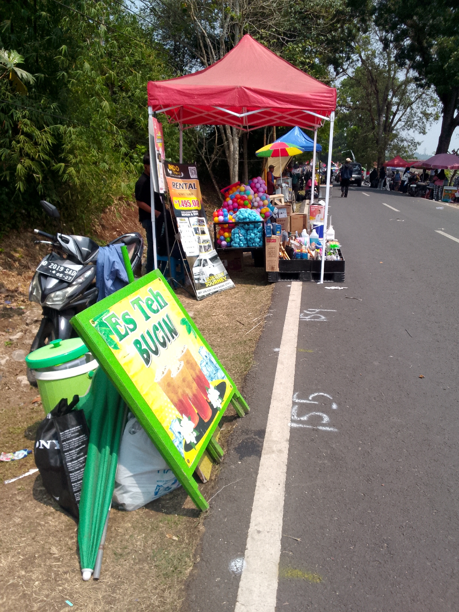 An outdoor roadside stall in Mandalawangi, Indonesia, is depicted. A red canopy covers a stall displaying various items, including colorful balls, boxes of goods, and bottles. A sign in front of the stall advertises "Es Teh BUCIN," which translates to "Obsessed Iced Tea." A parked motorcycle with a license plate reading "2019 SAD" is visible to the left of the stall. An umbrella and several bags are also present. The stall is situated beside a paved road, with trees and vegetation lining the background. The scene appears to be during daylight hours with a clear sky.