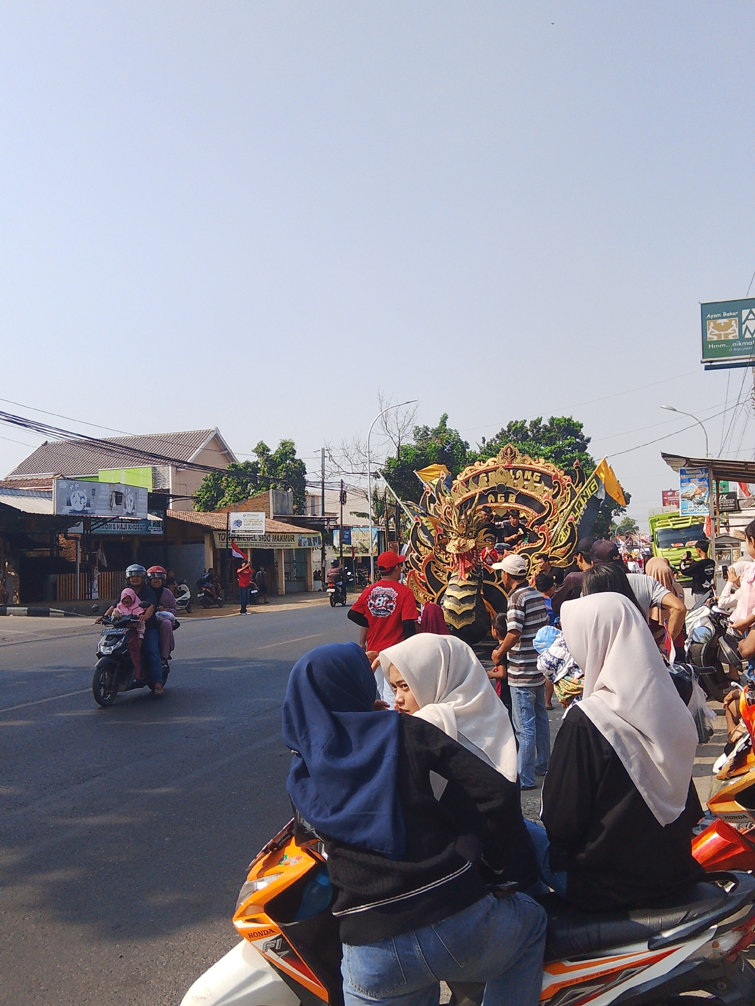 The image depicts a street scene in Rejosari, Indonesia, likely during a parade or festival. In the foreground, two women wearing hijabs are seated on a motorcycle. One woman, on the left, has a blue hijab and a black jacket over blue jeans. The other woman, on the right, wears a light-colored hijab and a black top. Behind them, a colorful float with a dragon motif is prominently displayed, adorned with gold and black details, and flags. Various people are gathered around the float, some watching and others participating in the event. A green truck is visible in the background, along with several buildings lining the street, suggesting a commercial or residential area. There are also other motorcycles and people moving along the street. The sky is clear and blue, indicating a sunny day.