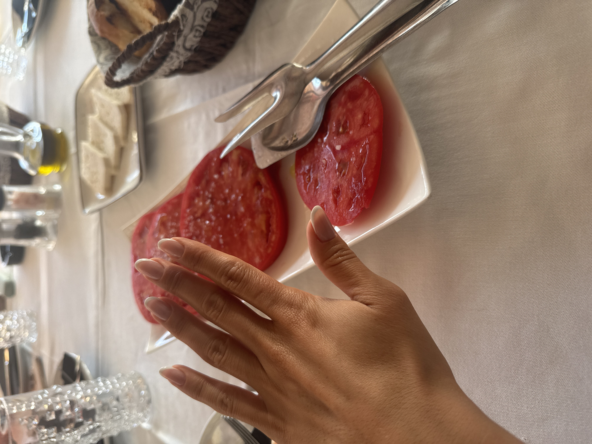 The image captures a top-down view of a dining table set with various food items and tableware, likely at a meal in Bigova, Montenegro. The central focus is a person's hand, exhibiting manicured fingernails with a French tip style, reaching towards a white rectangular dish holding several thick slices of red tomato. A silver serving fork and spoon are resting on the dish with the tomatoes.
In the background, to the upper left, a woven basket contains bread, partially visible. Below the basket, another small, rectangular white dish holds several small, pale, cube-shaped items, possibly cheese or butter. To the left, several glass bottles are visible, one of which appears to contain olive oil. Sparkling, decorative glassware, possibly tumblers or bottles, is also present on the left side of the frame. The entire table is covered with a crisp white tablecloth, which has a subtle textured pattern. The lighting suggests daytime. No discernible text or strong emotional cues are visible. The scene depicts a simple, fresh meal being prepared or served.