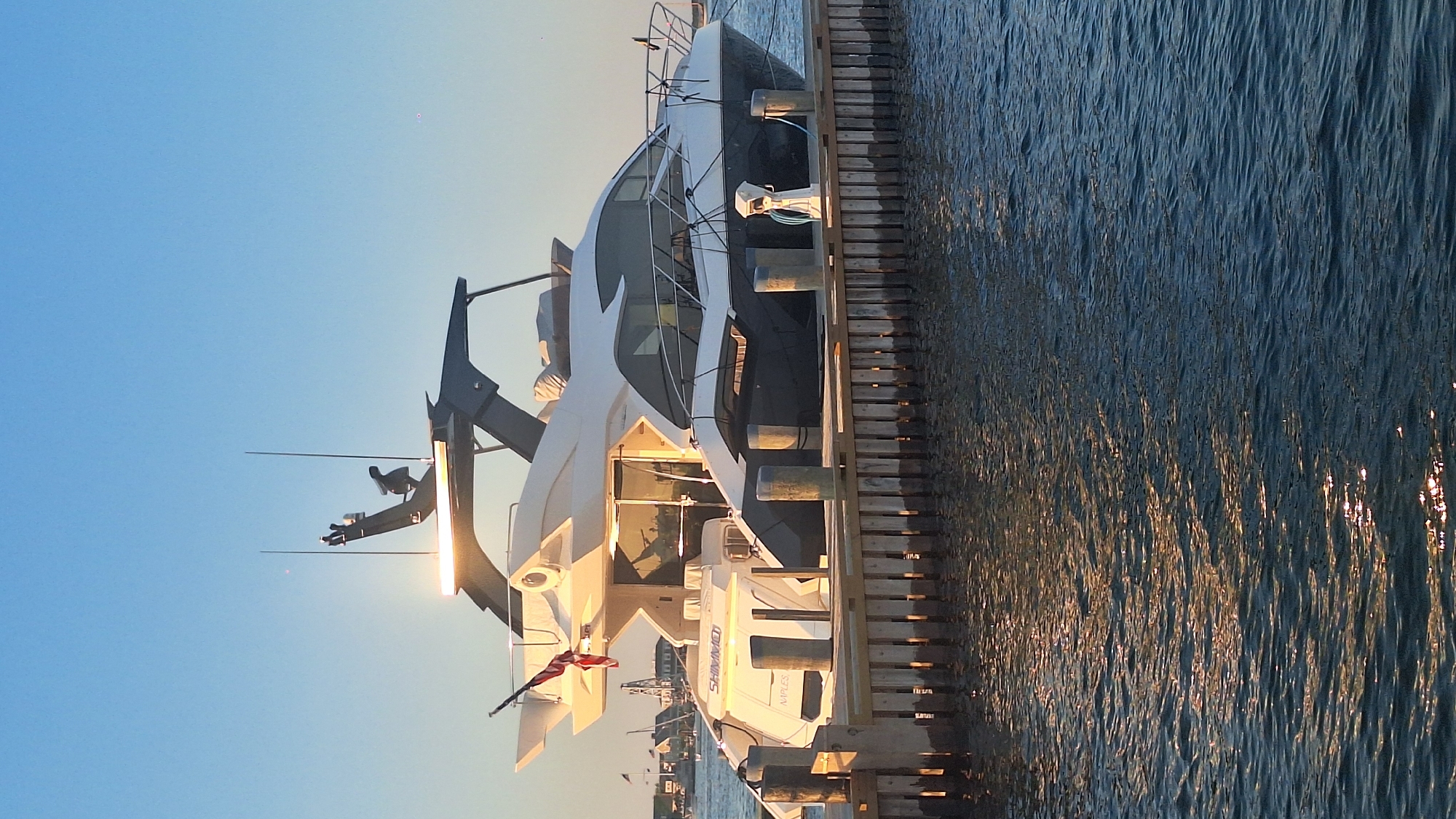 A large, modern yacht is docked at a wooden pier. The yacht is white and dark gray, with a sleek design. It has multiple levels and large windows. A red and white flag is flying from a pole on the stern of the yacht, and the name "SHIMMO" and "NAPLES" are visible on the back of the yacht. A fuel pump is visible on the deck of the yacht. The water is choppy and reflects the sunlight. In the background, there are other boats and buildings, suggesting a harbor or marina. The sky is clear and blue, with the sun setting, casting a warm glow on the scene. The overall impression is one of luxury and leisure. The location is identified as Nantucket, United States of America.