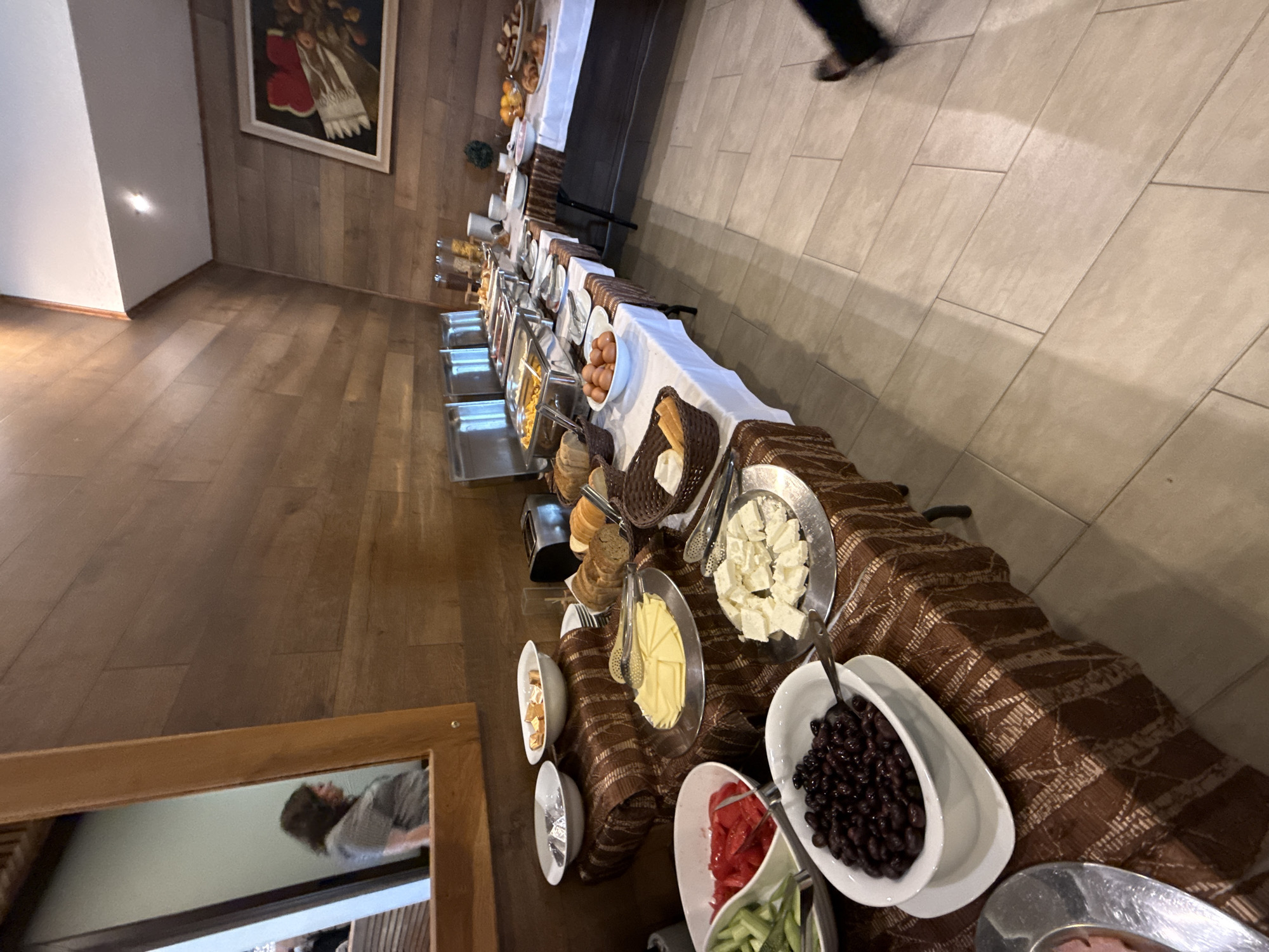 The image shows a hotel buffet in Veliko Tarnovo, Bulgaria. The room has wooden floors and paneling, with a large painting on the wall. A long table laden with a variety of food items forms the buffet, including: chafing dishes with cooked items, bowls of eggs, bread, butter, cheeses, olives, sliced tomatoes, cucumbers, and various other prepared dishes. There is also a person partially visible in the background, walking on a tiled floor. A framed mirror is visible in the foreground, reflecting a person. The buffet is set up with a decorative brown cloth draped over the table.