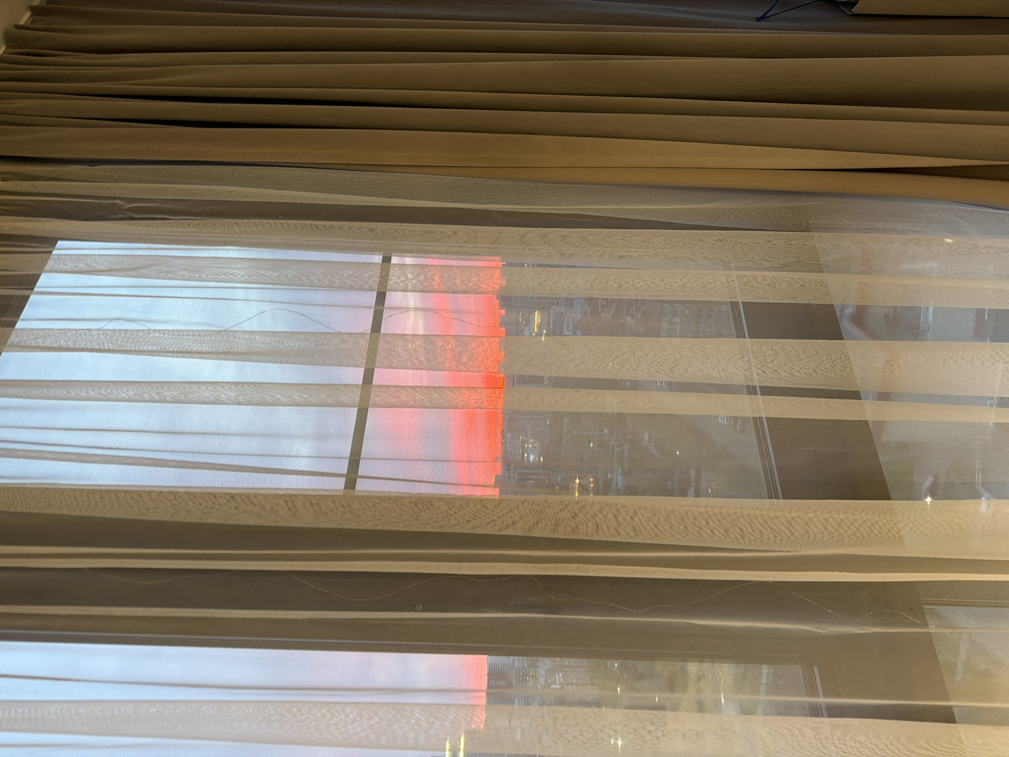 The image shows a view through a window with sheer blinds, possibly inside a building. The blinds have alternating opaque and translucent horizontal stripes. Outside the window, there's an urban scene, partially obscured by the blinds. A reddish glow, likely from the sunset or artificial lights, is visible through the window. The presence of buildings suggests an urban setting. It is not possible to determine the specific location in Krasnoyarsk, Russia, without further contextual information.