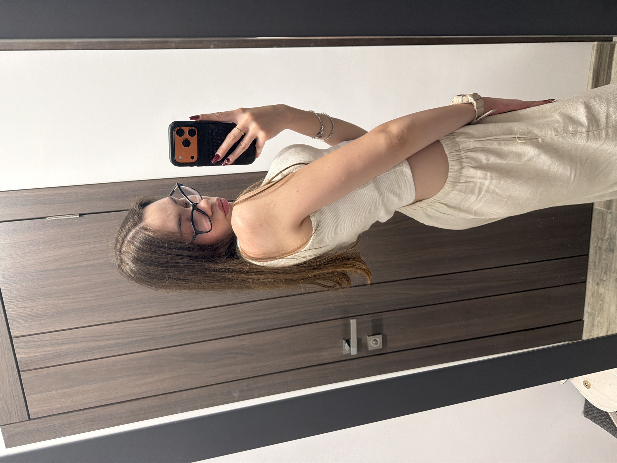 A young woman with long, brown hair and glasses takes a mirror selfie in what appears to be a fitting room or dressing area. She is wearing a white tank top and light-colored linen pants. She is holding a black phone with an orange case. In the background, there is a wooden door with a silver handle and a white wall. A portion of a light-colored bag and a dark gray frame are visible in the foreground. The scene is likely in Varna, Bulgaria, given the context provided.