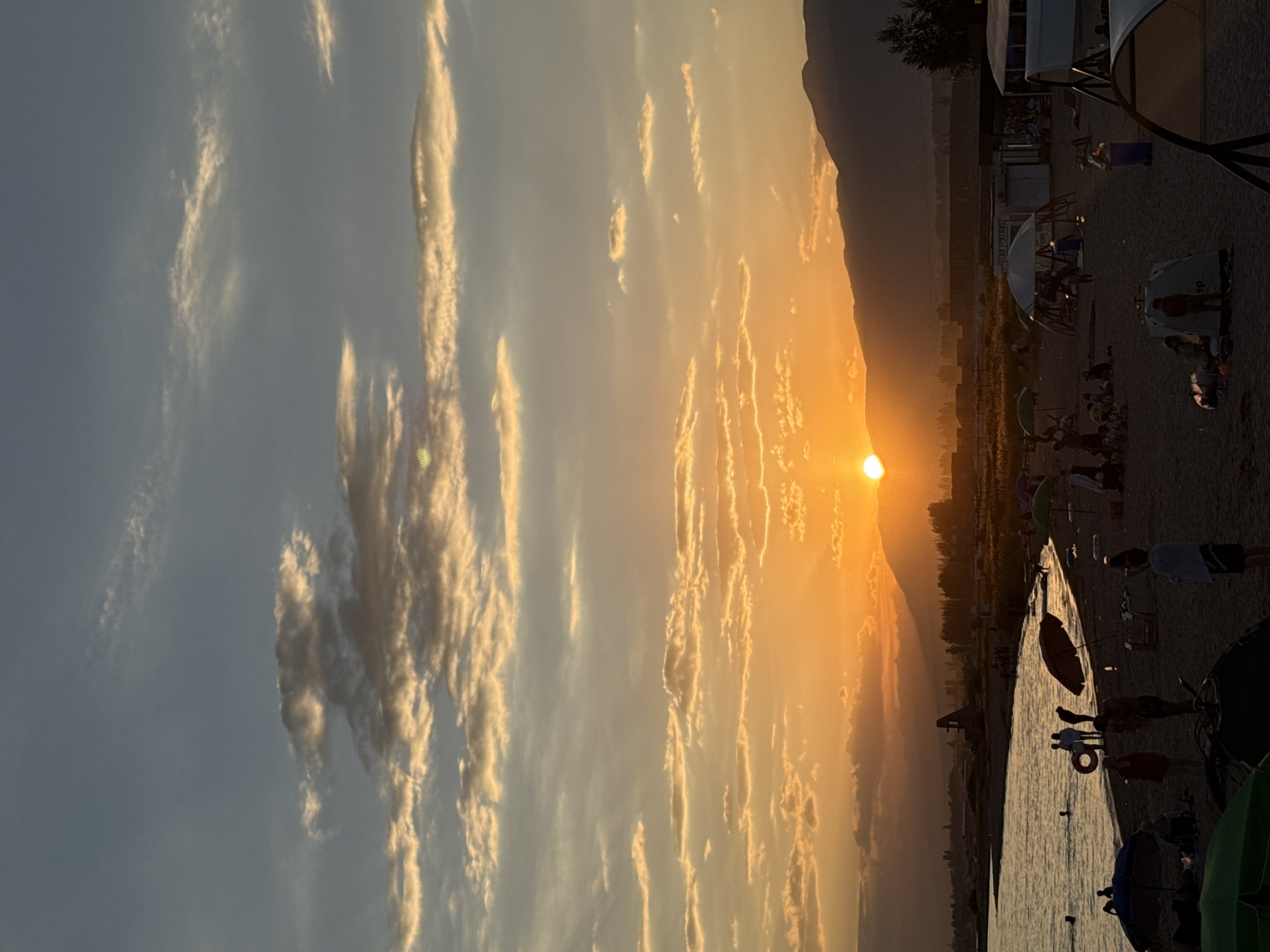The image depicts a serene sunset scene at a beach in Sary-Oy, Kyrgyzstan. The sun, a bright orange orb, is positioned low on the horizon, casting a warm glow across the sky and illuminating the scattered clouds with golden hues. In the foreground, a sandy beach is dotted with various beachgoers, some relaxing under umbrellas, others wading in the calm waters of what appears to be Issyk-Kul Lake. Several tents are set up on the sand, suggesting a leisurely stay. In the background, a range of silhouetted mountains forms a majestic backdrop against the vibrant sky. The overall atmosphere is tranquil, with the fading light of day creating a picturesque and peaceful setting.