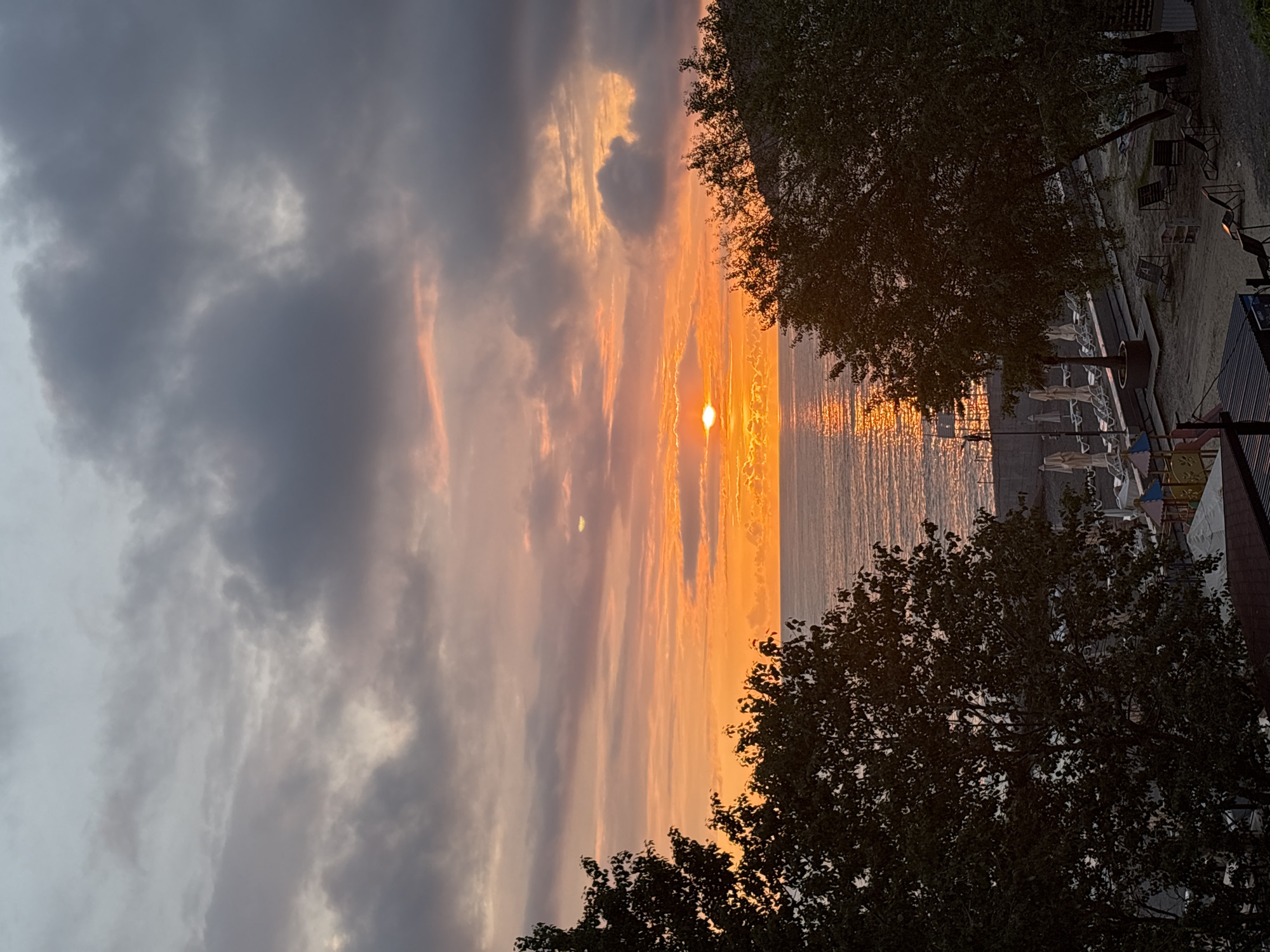 The image shows a sunset view over the Black Sea in Sochi, Russia. The sky is a mix of dark, cloudy formations on the left and bright orange and yellow hues near the horizon, where the sun is setting. The sun reflects on the water's surface. Along the coastline, on the right side of the image, there are trees, benches, and sun umbrellas.