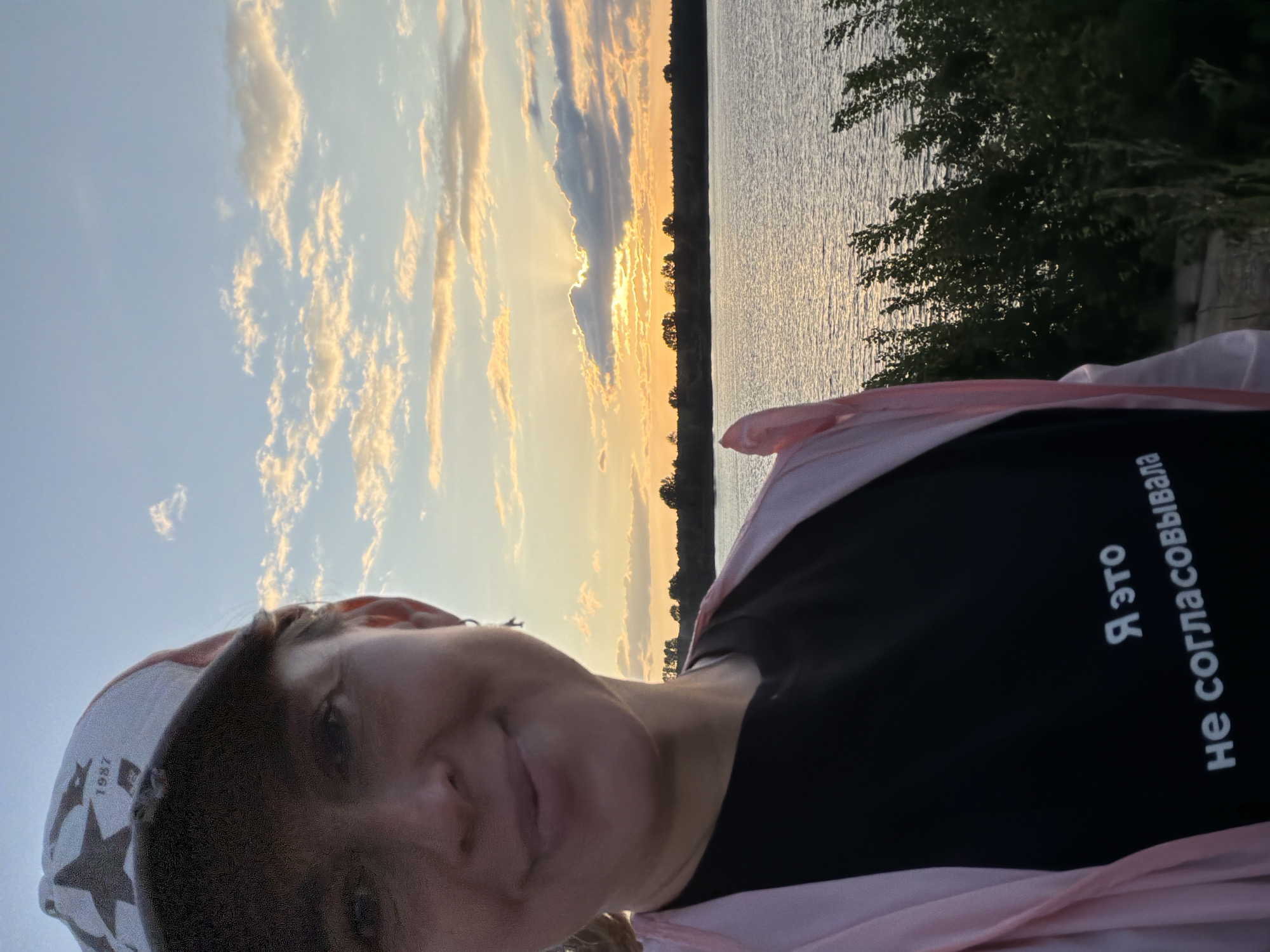 A woman with a baseball cap and a pink jacket is taking a selfie by a body of water during sunset. The sky has colorful clouds. A dark t-shirt with white Russian text is visible on her chest. The text reads "Я это не согласовывала" which translates to "I did not agree to this". The location is likely in the city of Chaikovsky, Russia, as suggested by the context.