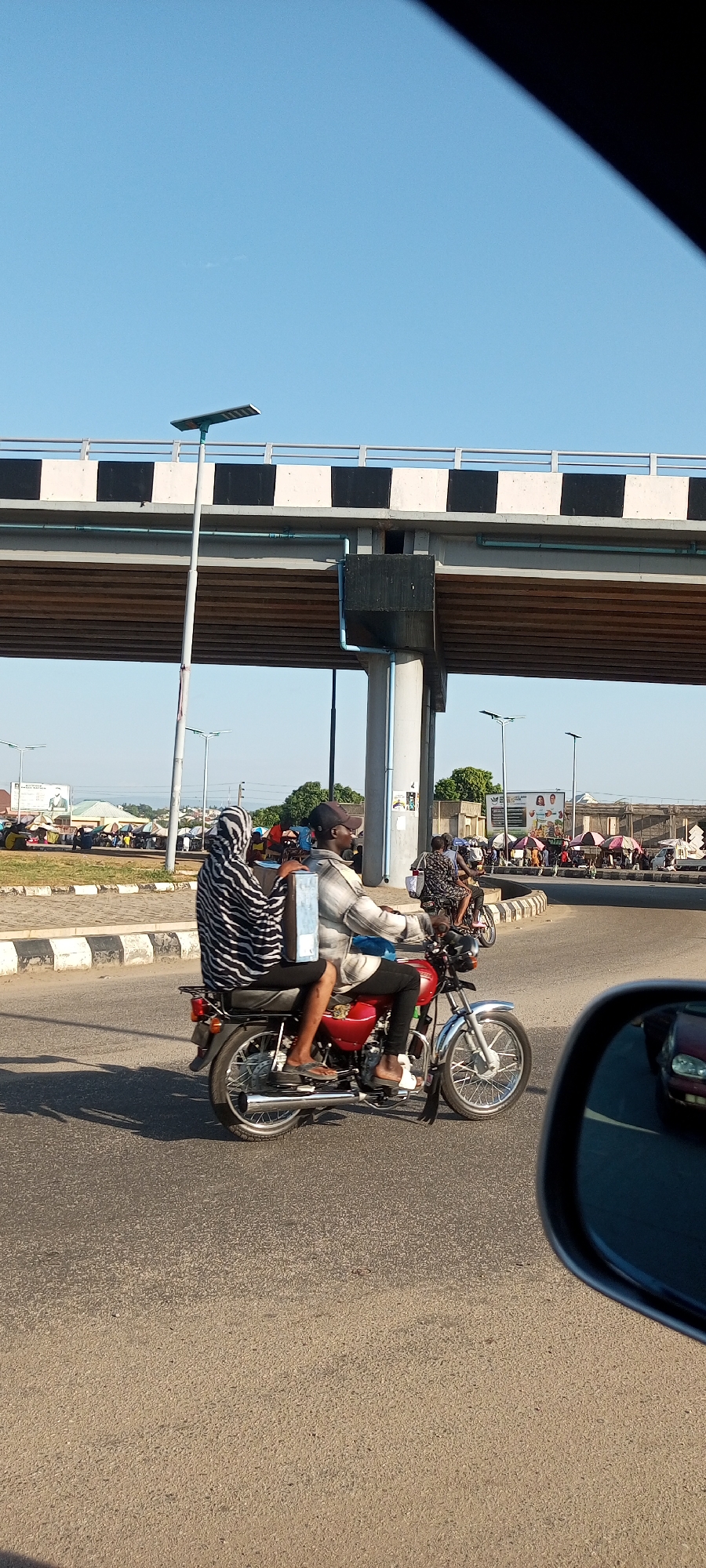The image captures a vibrant urban scene in Lokoja, Nigeria, featuring a man and woman on a motorcycle under a concrete overpass.
In the foreground, a man wearing a plaid shirt, dark trousers, and a cap drives a red motorcycle with chrome accents. A woman rides as his passenger, clad in a black and white striped garment, and appears to be carrying a light blue box-like item on her back. The motorcycle is on a paved road, partially shadowed, suggesting bright sunlight. Part of a car's side mirror is visible in the bottom right corner, indicating the photo was taken from inside another vehicle.
Above them, a large overpass or bridge spans the road, characterized by sturdy concrete structures and railings painted with distinctive black and white alternating stripes. A tall streetlamp with a solar panel on top stands near one of the overpass pillars.
In the background, the scene opens to a busy street and market area. Numerous people are visible, some on other motorcycles or walking. Market stalls with colorful umbrellas and canopies line the roadside. A billboard in the mid-distance displays partial text including "DO & MO..." and "BE HEALTHY EAT GOOD," alongside images of two people and food items. The sky is a clear, bright blue, indicating good weather. The overall setting is a bustling street in a metropolitan area of Lokoja, Nigeria, likely during the day, given the strong shadows and clear sky.