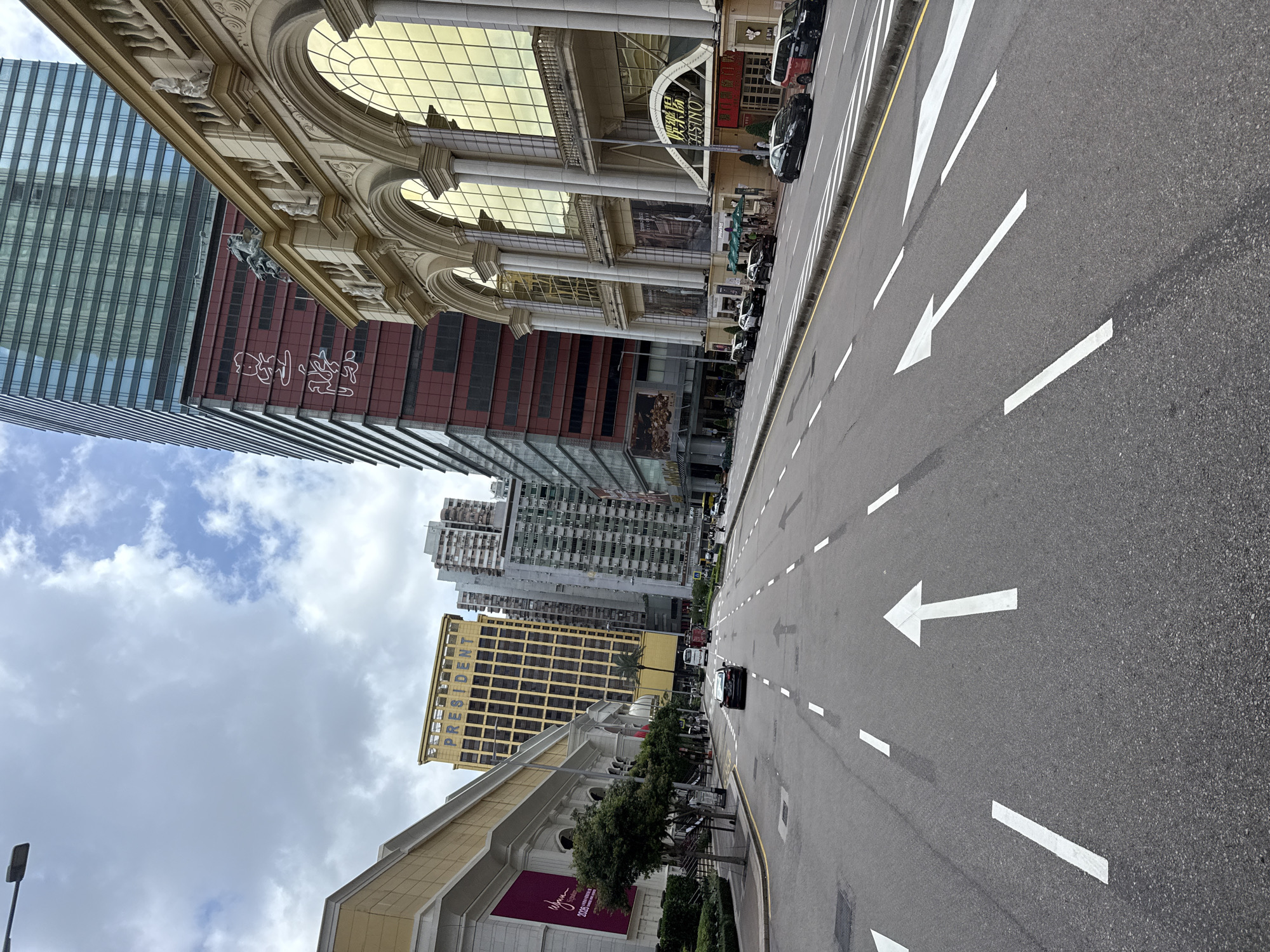This image captures a street scene in Macau, China, likely in the Cotai Strip area, characterized by its modern architecture and bustling urban environment. The foreground shows a wide asphalt road with clear white lane markings and directional arrows indicating traffic flow. Several cars are visible on the road, including black sedans and a red SUV, suggesting active traffic.
Flanking the road are imposing buildings that define the cityscape. On the left, a building with a large purple banner and a gold-accented facade, possibly a hotel or casino, stands prominently. Adjacent to it, a distinct yellow building with the word "PRESIDENT" in large blue letters on its upper section is visible. Further down the street, a row of high-rise buildings with varying architectural styles and colors suggests a dense urban development.
On the right side of the street, a grand building with a classical facade, featuring arched windows with golden panes and ornate decorative elements, dominates the view. A prominent sign with Chinese characters and the word "CASINO" in English is displayed above the entrance, confirming its function. This building's opulent design stands in contrast to the more modern, glass-faced skyscrapers seen further in the background.
The sky above is partly cloudy, with patches of blue sky visible amidst the grey clouds. The overall impression is one of a vibrant and dynamic city, showcasing a mix of architectural styles and a busy, modern street life. The presence of multiple casinos and large hotel complexes points towards Macau's well-known identity as a global entertainment and gambling hub.