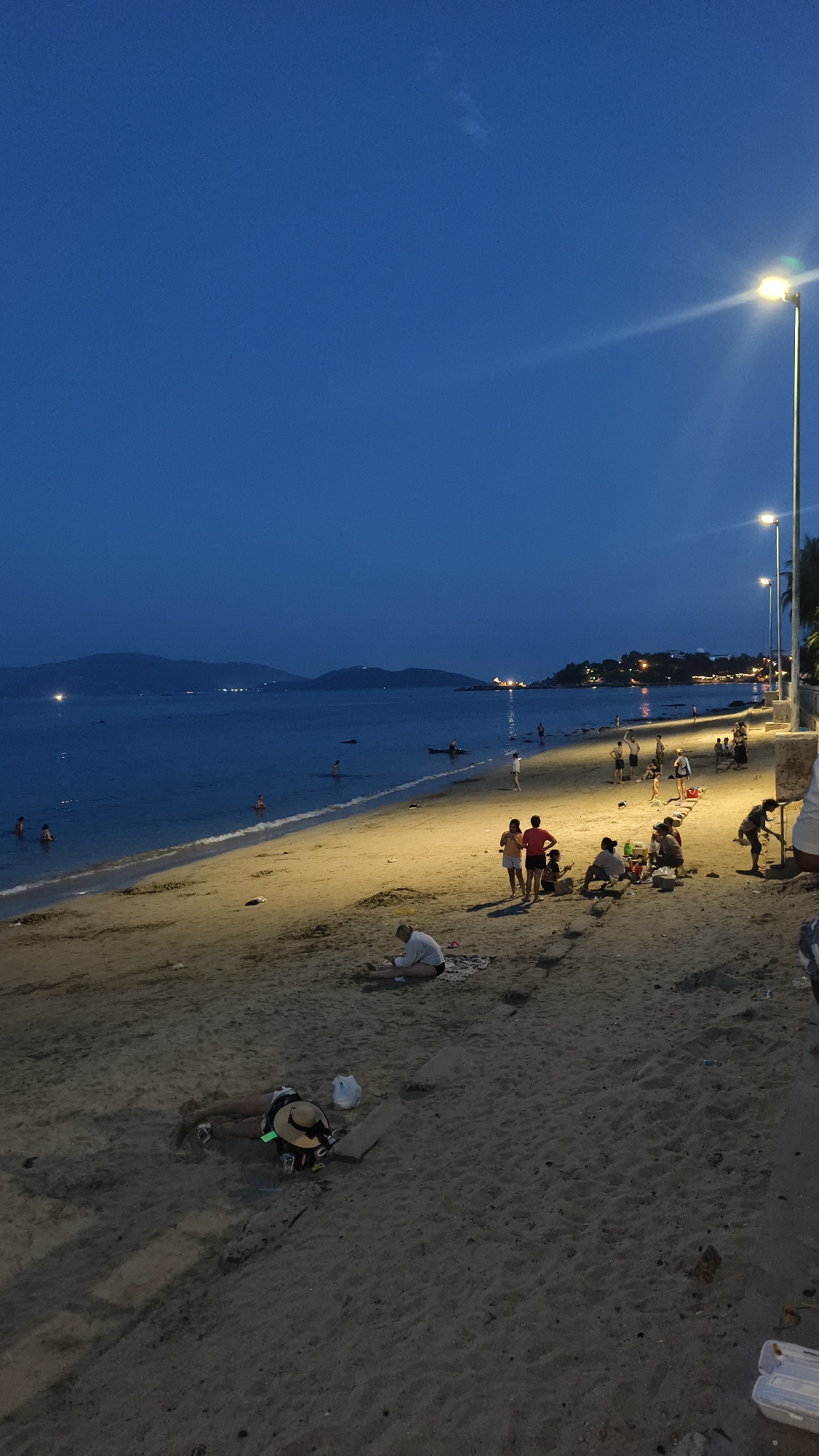 An eye-level, medium shot captures a beach scene at dusk in Unknown, Unknown. The sky is a deep, clear blue, transitioning to darker hues as it meets the horizon, where faint hills are silhouetted against the fading light. Several lampposts with bright lights illuminate sections of the sandy beach and the edge of the ocean.
On the sandy shore, a group of people are engaged in various activities. Near the water's edge, a few individuals are wading or swimming. Further up the beach, a group of approximately four people, including a man and two women, are standing together. To their right, several more individuals are gathered, some sitting on blankets, appearing to picnic or socialize. In the foreground, a person wearing a straw hat lies on their stomach on a blanket, facing away from the camera. Another person is seated further back on the beach, also on a blanket.
The ocean is calm, with gentle waves lapping at the shore. A few scattered pieces of trash are visible on the sand, including a white plastic container in the lower right corner. The overall atmosphere is relaxed and serene, with the artificial lighting creating pools of warmth on the beach.