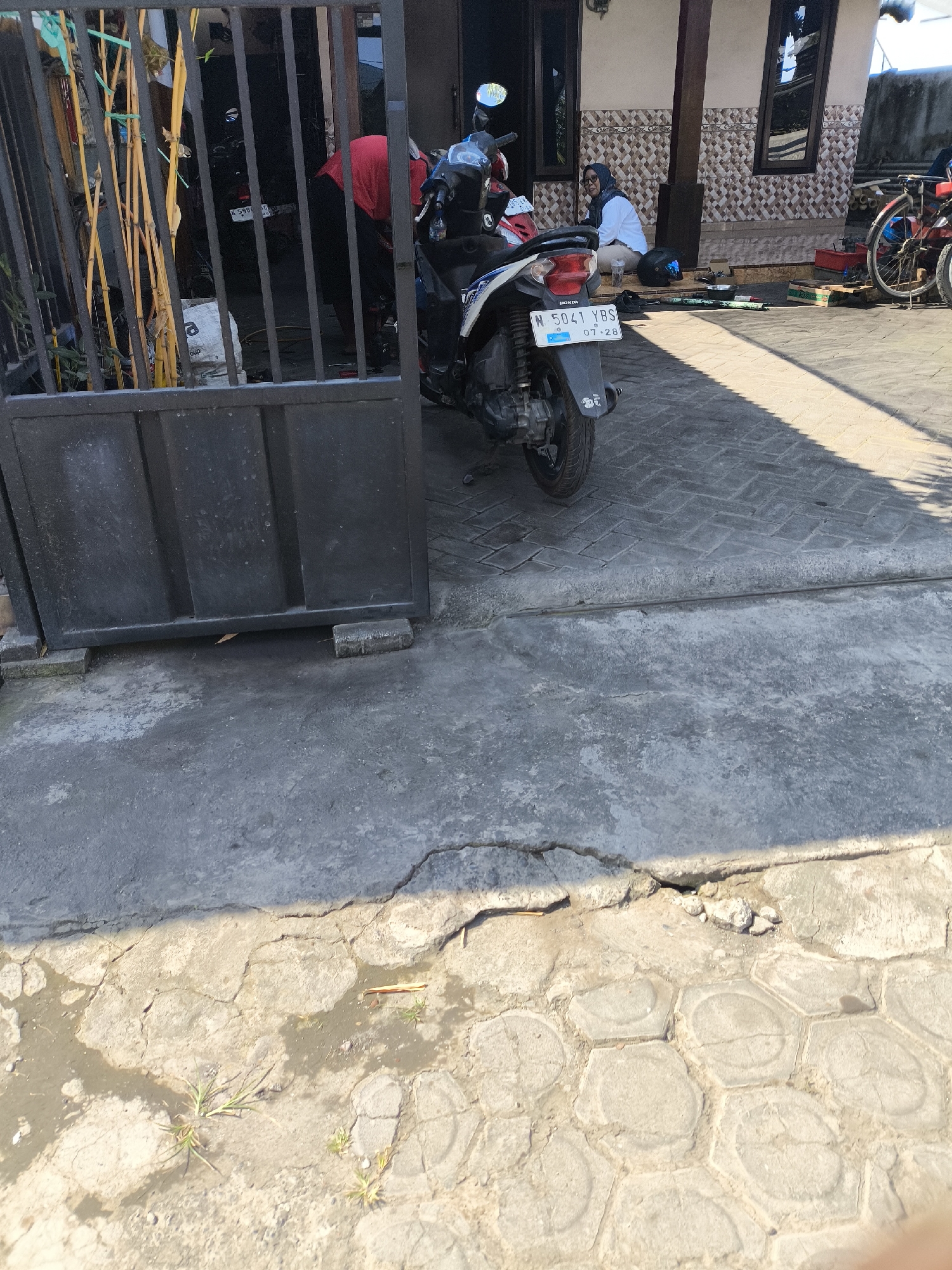 The image captures a scene in front of a building on a sunny day in Lumajang, Indonesia. In the foreground, a dark metal gate stands open, revealing a glimpse into a yard. Beyond the gate, a person wearing a red shirt and dark pants is seen tending to a motorcycle. The motorcycle, a white and blue model with license plate "N 5041 YBS," is parked on a paved surface. Further back, a woman in a white shirt and blue headscarf is seated on the steps of the building, which features a patterned tiled facade. To the right, a red bicycle with a metal frame is visible. The ground in the foreground consists of cracked concrete and hexagonal paving stones, with some patches of standing water. The overall impression is one of a residential or commercial area on a typical day.