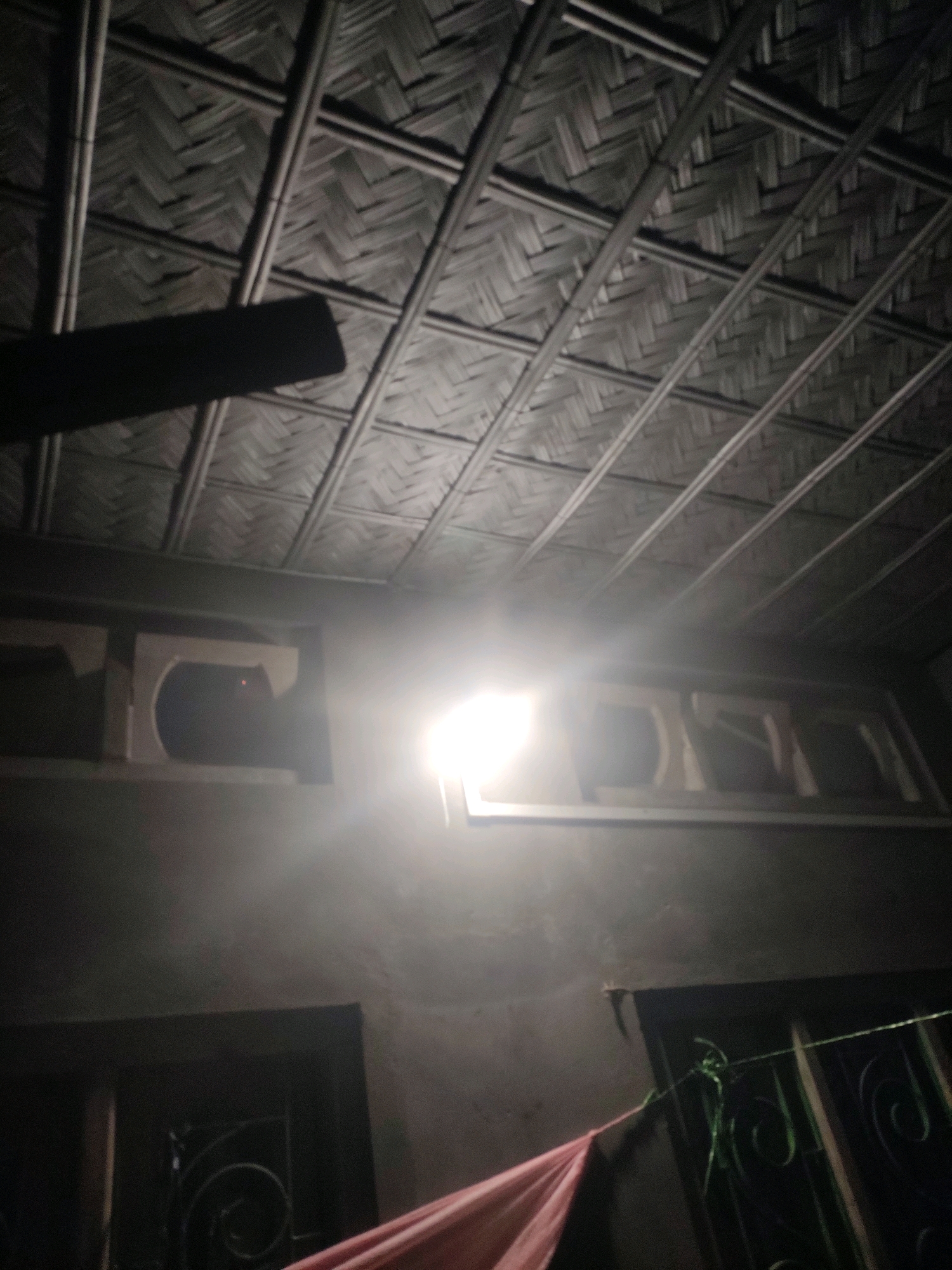 The image depicts an indoor scene with a woven bamboo ceiling. A bright light source, possibly a lamp, is prominently positioned in the center of the frame, illuminating the area. Below the ceiling, there are two rectangular openings, likely windows or alcoves, with visible wooden structures inside. The walls appear to be constructed of concrete or plaster. In the lower part of the image, a dark doorway with an ornate metal grate is visible, and a pink cloth drapes across the foreground. The overall impression is of a simple, rustic interior illuminated by artificial light.