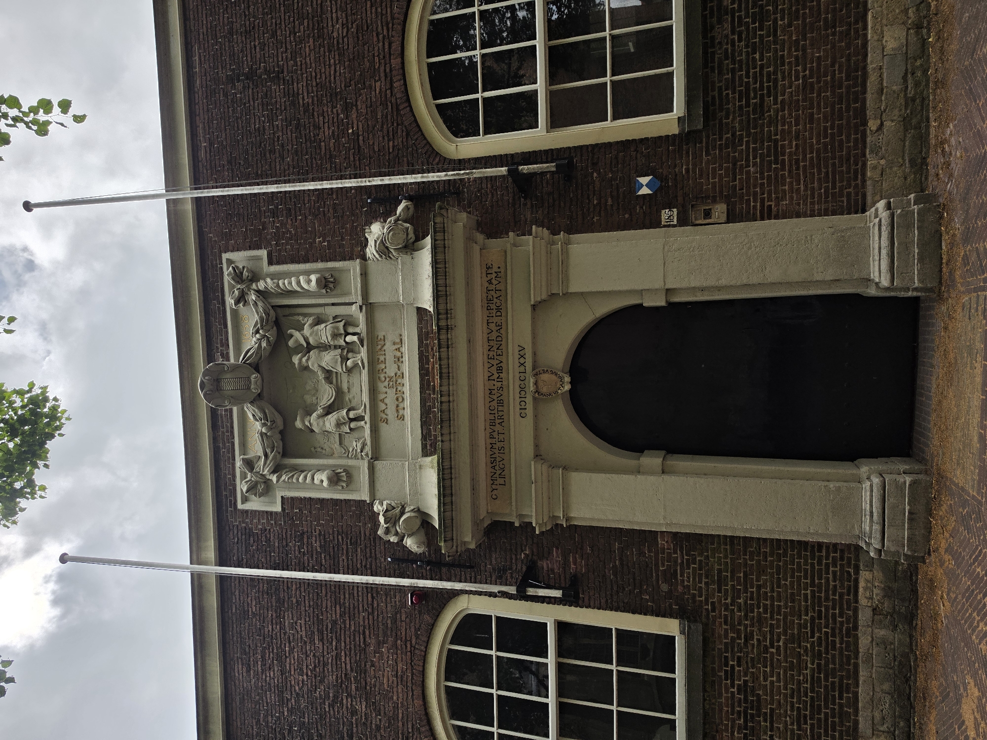 The image shows the entrance to the "Gravensteen" building, a former gymnasium located in Delft, Netherlands. The building's facade is constructed of dark red brick. Above the arched doorway, there is a stone relief depicting children and an inscription in Latin. Flanking the entrance are two tall windows with white frames and mullions. Two flagpoles are visible, one on each side of the relief, extending upwards against a cloudy sky. The entrance itself is a large, dark, arched door set within a stone archway supported by a large pillar. A house number "185" is visible to the right of the entrance, along with a blue and white shield. The ground in front of the building is paved with reddish-brown bricks.