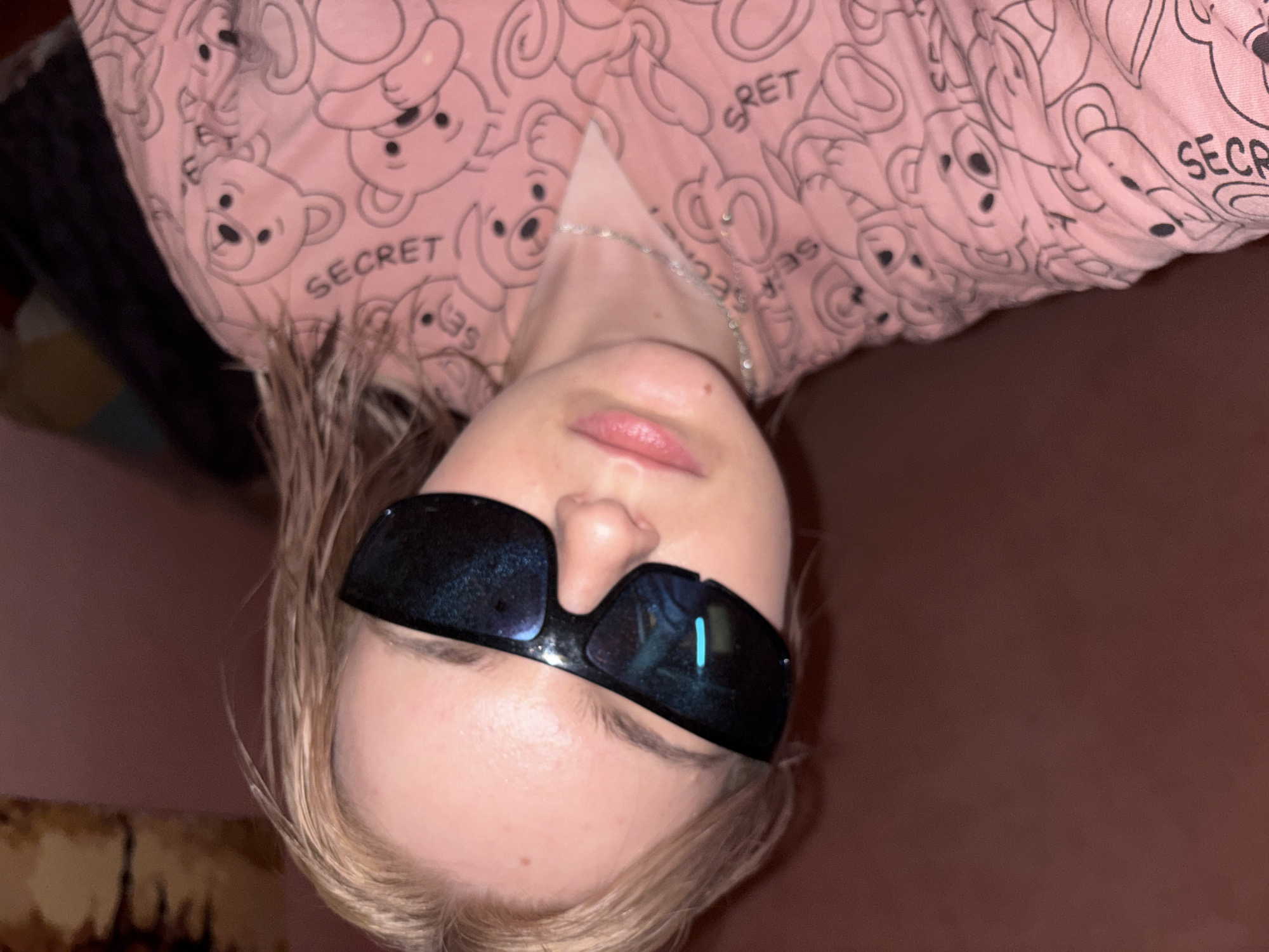 The image presents a close-up, upside-down portrait of a person, likely young, with fair skin and light blonde hair. The individual is wearing dark, reflective sunglasses that completely obscure their eyes. Their lips are slightly parted and appear a natural light pink.
The person is dressed in a pink long-sleeved top or pajamas, which features a repeating pattern of cartoon teddy bears. The word "SECRET" is clearly visible multiple times within this pattern, written in black capital letters. A thin silver chain necklace is also visible around the subject's neck.
The background is predominantly a dark, indistinct brown or maroon, suggesting an indoor setting with low lighting. On the left side of the frame, there are vague hints of a textured or patterned surface, possibly a piece of furniture or another item in the room. The overall dim lighting suggests the photograph was taken during the evening or in a dimly lit space. There is no specific activity taking place other than the person posing for the camera, presented in an inverted orientation. Emotions are not discernible due to the sunglasses. While the image itself does not provide explicit location cues, the context states the location is unknown, Russia.
