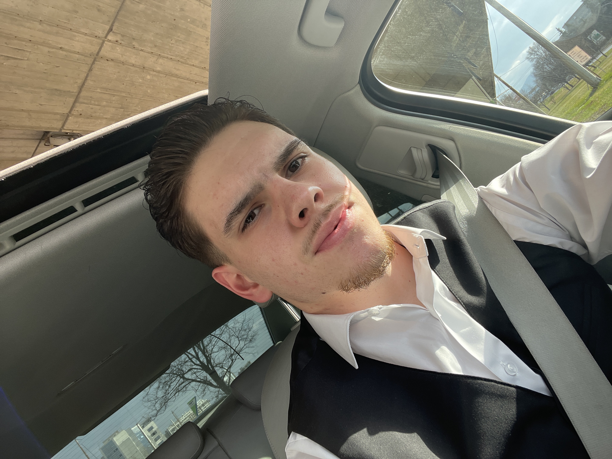 A young adult male is depicted seated in the front passenger seat of a vehicle, viewed from a slightly low angle, indicating a self-portrait. He has dark, neatly styled hair and short facial hair, including a goatee and mustache. A light-colored scar or mark is visible extending from the left corner of his mouth towards his cheek. He is wearing a white collared shirt, open at the top, beneath a dark, likely black, waistcoat or vest. A light grey seatbelt is properly fastened across his chest and lap. He looks directly at the camera with a subtle smirk.
The vehicle's interior features a grey headliner and trim. Part of a grey car seat is visible behind the subject. Through the upper section of the rear window, reflections of a modern building with multiple windows are visible, alongside bare deciduous tree branches. Above the vehicle, through what appears to be a sunroof or windshield, a large, textured concrete structure, consistent with the underside of an overpass or bridge, is prominent. This concrete exhibits vertical lines from its formwork. Through the passenger side window on the right, an outdoor urban scene is visible under bright sunlight, showing a clear blue sky, a grassy area with bare trees, and distant low-rise buildings. A utility pole is also visible. The lighting casts distinct shadows within the vehicle.
This scene is located in an urban area of Sofia, Bulgaria, specifically beneath or in close proximity to a large concrete overpass, characteristic of major boulevards or highway intersections within the city.