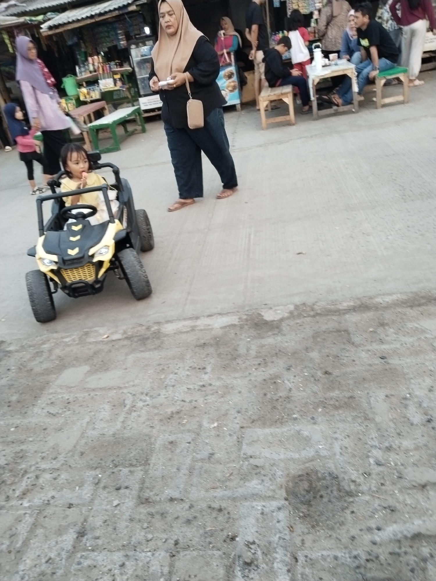 The image captures a scene in the city of Serang, Indonesia, likely near a street market or food stall area. In the foreground, a young child sits in a yellow toy ATV, holding a light yellow lollipop. Behind the child, an adult woman wearing a light brown hijab and a dark top walks away from the camera, holding a mobile phone and a small beige purse. To the left of the child, another woman in a purple hijab and a light-colored patterned outfit is partially visible, with a younger child in a pink shirt and blue pants standing next to her. Several other individuals and market stalls are visible in the background, suggesting a busy public space. The ground is paved with concrete and appears slightly worn. The lighting indicates it is likely daytime.