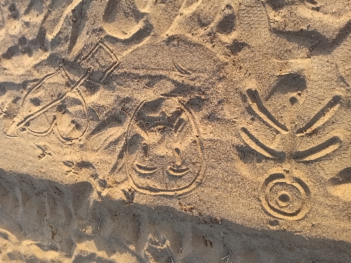The image depicts sand art of varying complexity. A spade symbol is drawn with a square-shaped top. A simple cartoon face features large eyes and a small, curved smile. A circular target-like design has concentric circles and several lines radiating outwards. The composition includes small footprints, suggesting animal activity. The sandy surface is textured and lit with a warm tone, indicating sunlight.