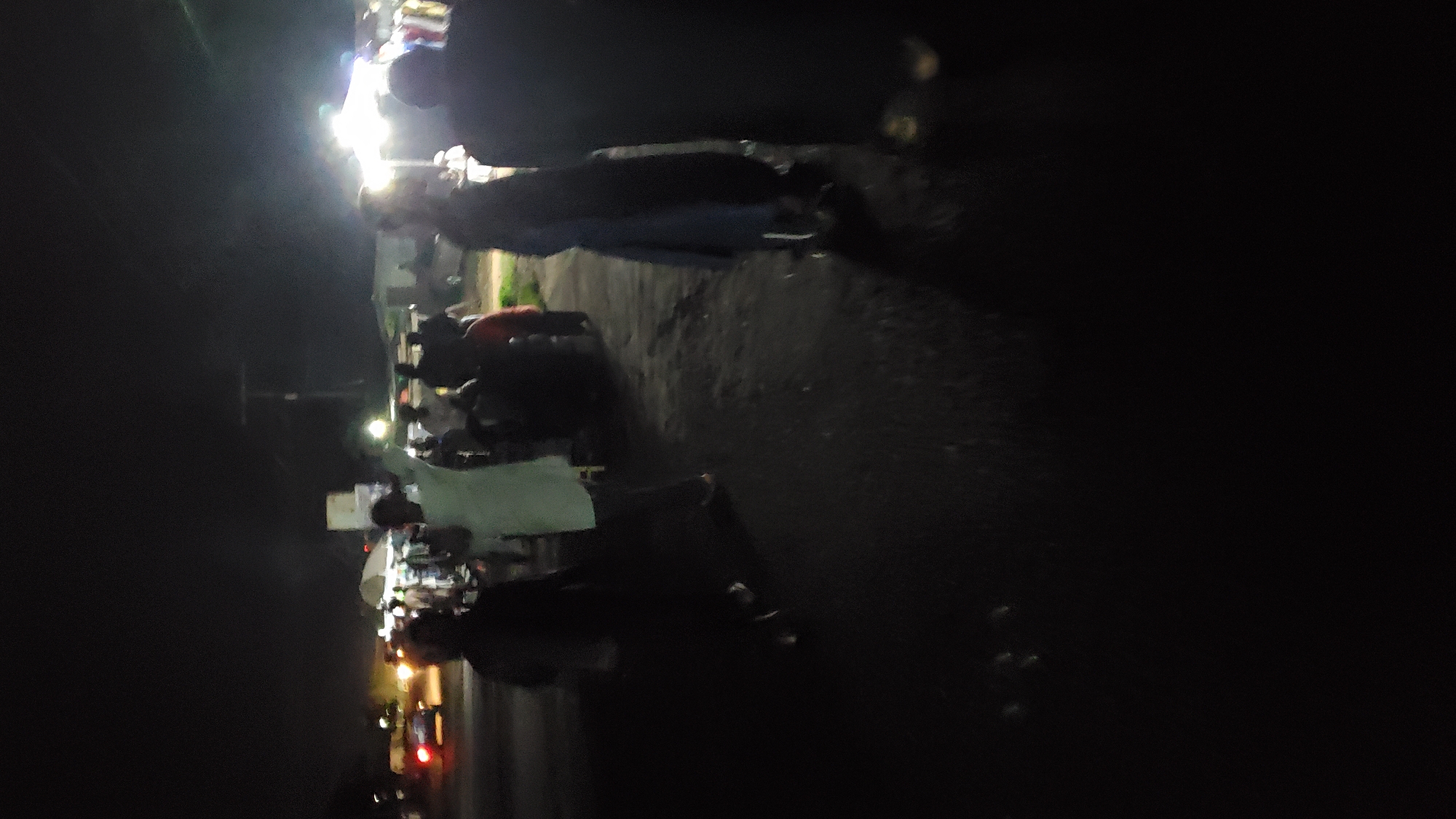 At night in Jalingo, Nigeria, a group of people are gathered on an unpaved road near an outdoor market or event. Several individuals are standing and walking, some illuminated by artificial lights from tents and vehicles. A car with its headlights on is visible in the background. The scene is dark, with limited visibility beyond the immediate area.