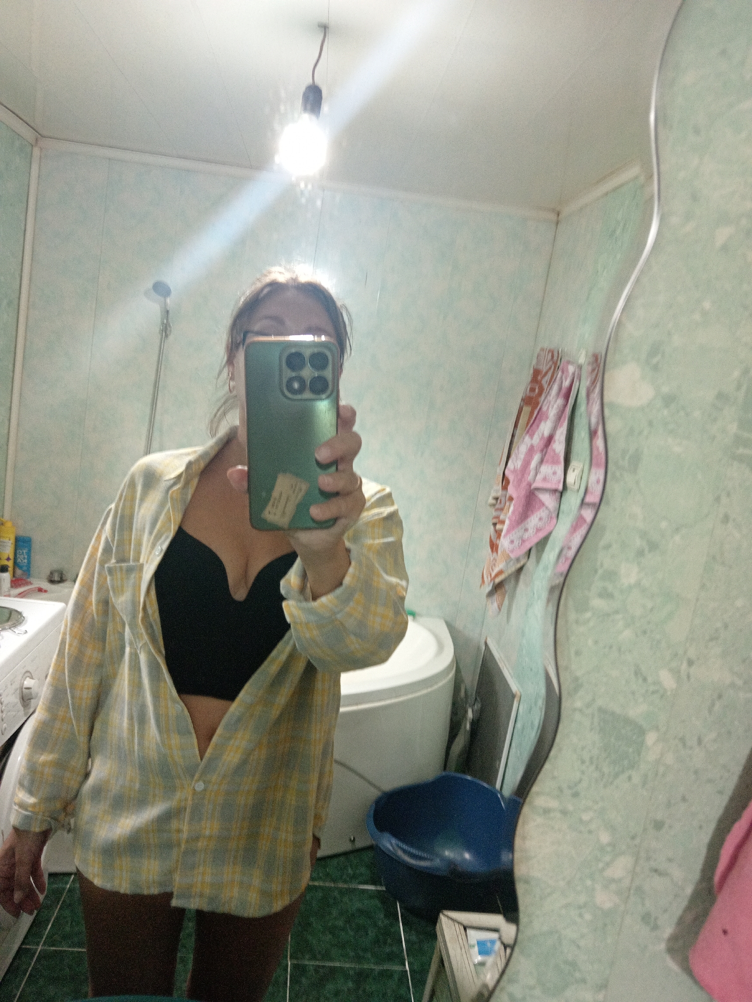 A woman is taking a mirror selfie in a bathroom. She is wearing a yellow and white plaid shirt unbuttoned over a black bra and dark shorts. Her left hand holds a green smartphone to take the picture. The bathroom has light green patterned walls, a white toilet, a washing machine, and a large, wavy mirror. A lightbulb hangs from the ceiling. A blue basin is on the floor. A pink and white patterned towel hangs on the wall. The image appears to be taken in Novopokrovka, Russia.