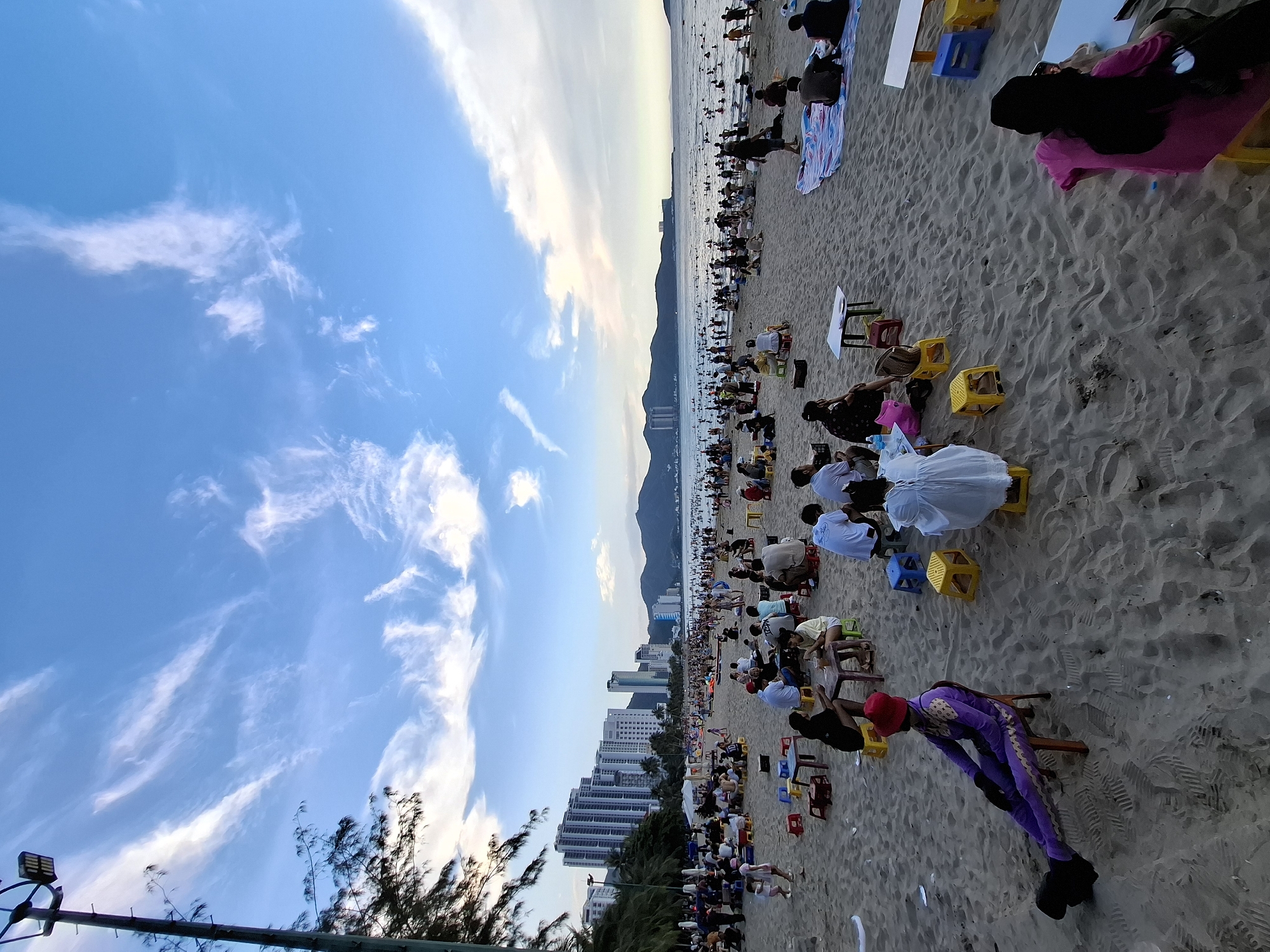 The image depicts a crowded beach in Bac Nha Trang Ward, Vietnam, under a partly cloudy blue sky. Numerous people are scattered across the sandy shore, many seated at small tables and chairs, suggesting a dining or social gathering. Some individuals are wading or swimming in the ocean, while others relax on the beach. In the background, a cityscape of tall buildings lines the coast, with a mountain range visible in the distance. The scene appears to be a vibrant public space during daytime.