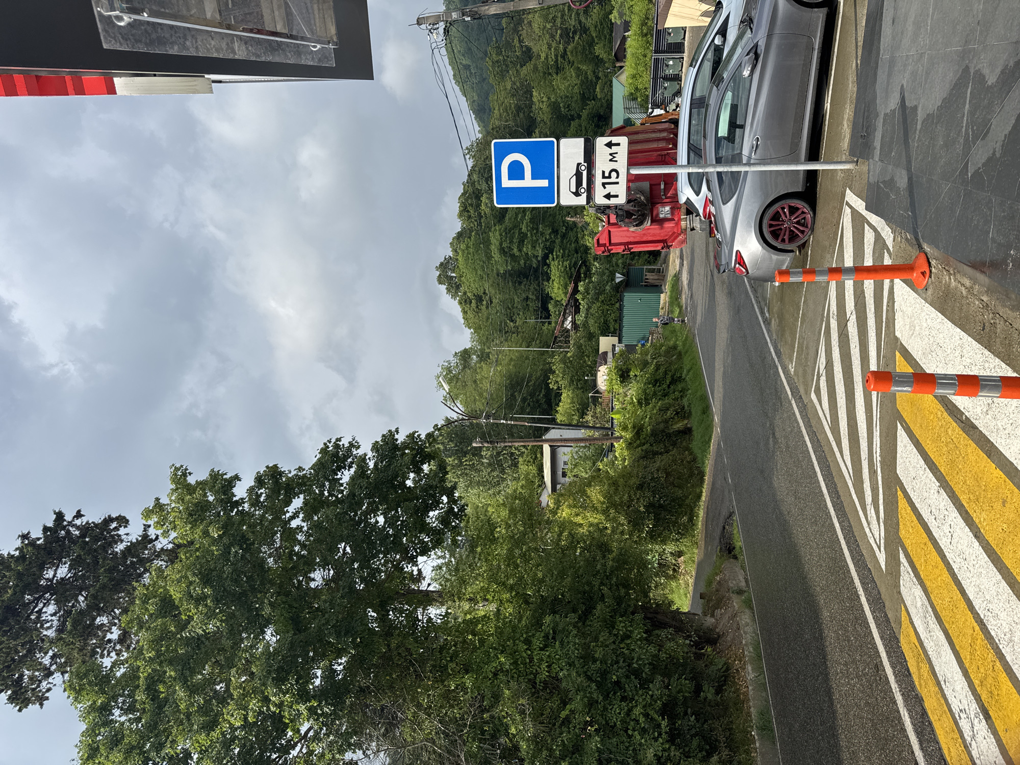 The image shows a street scene with a parking sign, a garbage truck, and cars. The parking sign is blue and white with a "P" symbol, indicating a parking area. Below it, there is a sign with a car symbol and an arrow pointing upwards, along with the text "15 m" which might indicate a distance or a specific parking instruction. A large red garbage truck is parked on the right side of the road, partially obscuring a silver car and a white car parked behind it. In the foreground, there is a pedestrian crossing with yellow and white stripes. The environment appears to be somewhat rural or suburban, with trees and vegetation visible in the background. There are also some buildings and utility poles. The sky is cloudy.