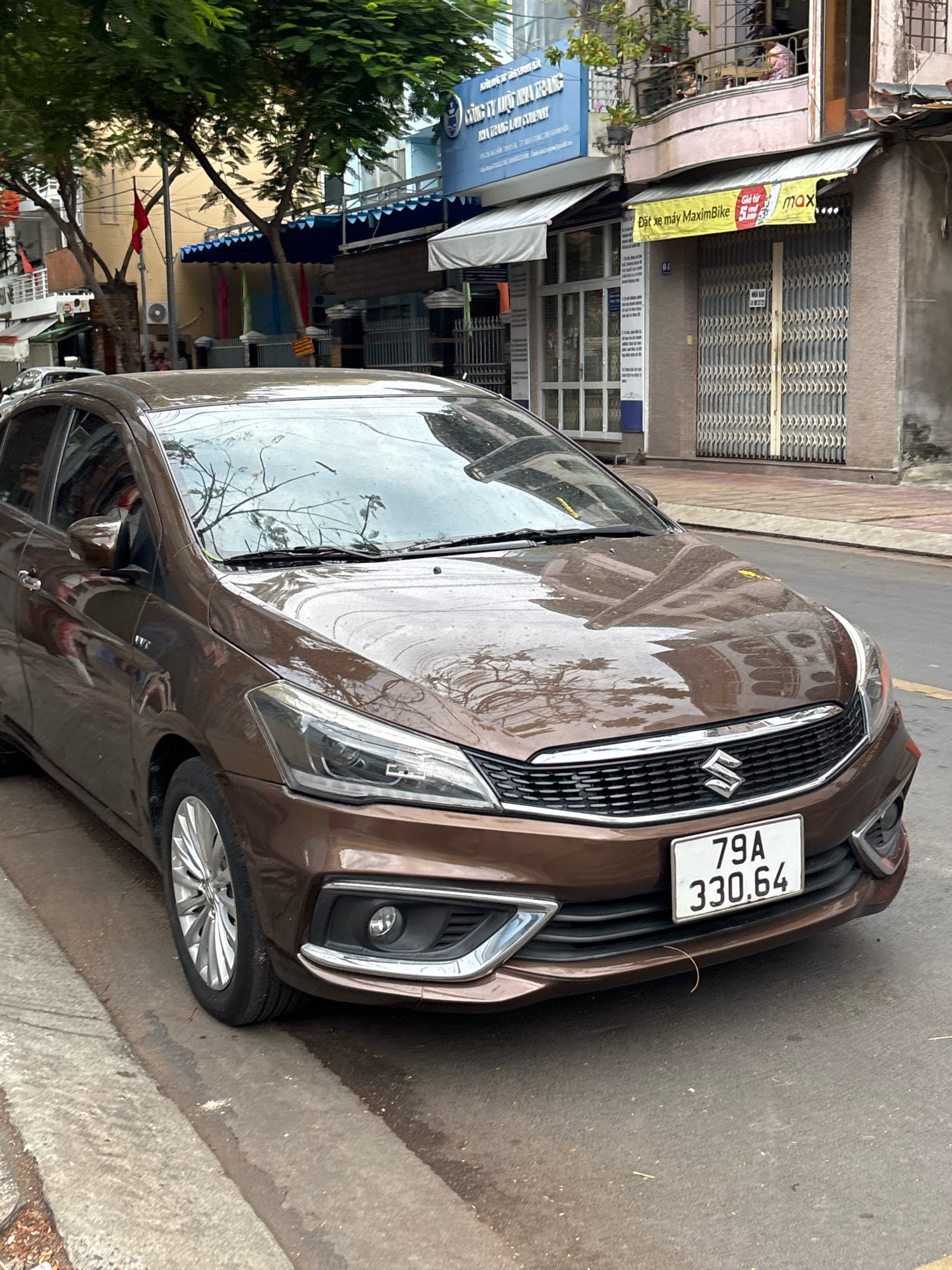 A brown Suzuki sedan is parked on a street in Nha Trang, Vietnam, with its front end facing the camera. The car has a license plate reading "79A 330.64". The street appears wet, suggesting recent rain. Buildings line the street, some with awnings and signs. A Vietnamese flag is visible on a pole attached to one of the buildings. The car's hood reflects the surrounding environment, including trees and buildings. There are no people clearly visible in the immediate vicinity of the car. The scene depicts a typical urban street in Vietnam.