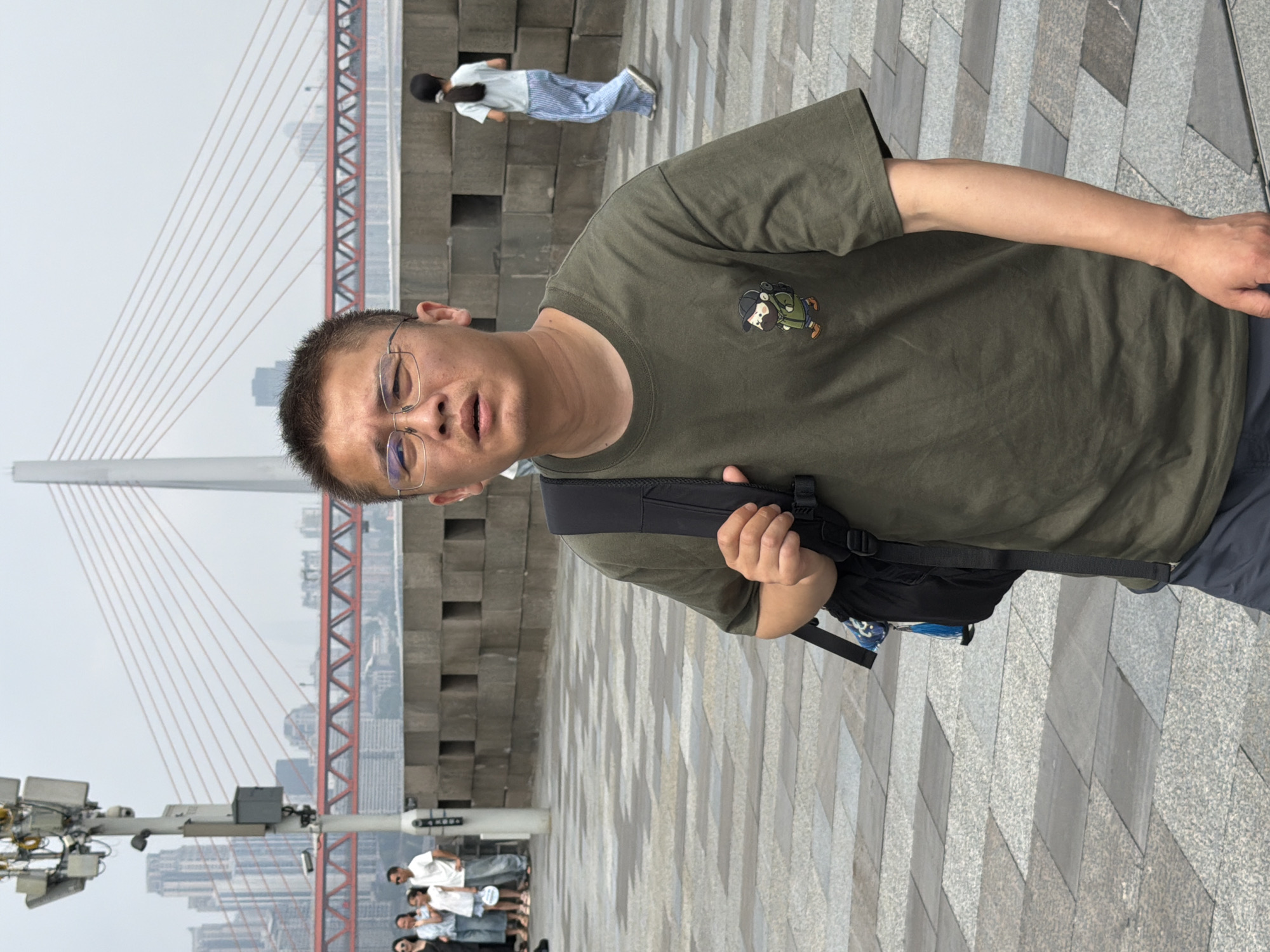 A man wearing a green t-shirt with a cartoon graphic, glasses, and a backpack is standing on a paved walkway. In the background, a bridge with red structural elements and white cables spans a body of water. A group of people, including children, are visible further back on the walkway. The environment appears to be an urban setting with buildings in the distance.